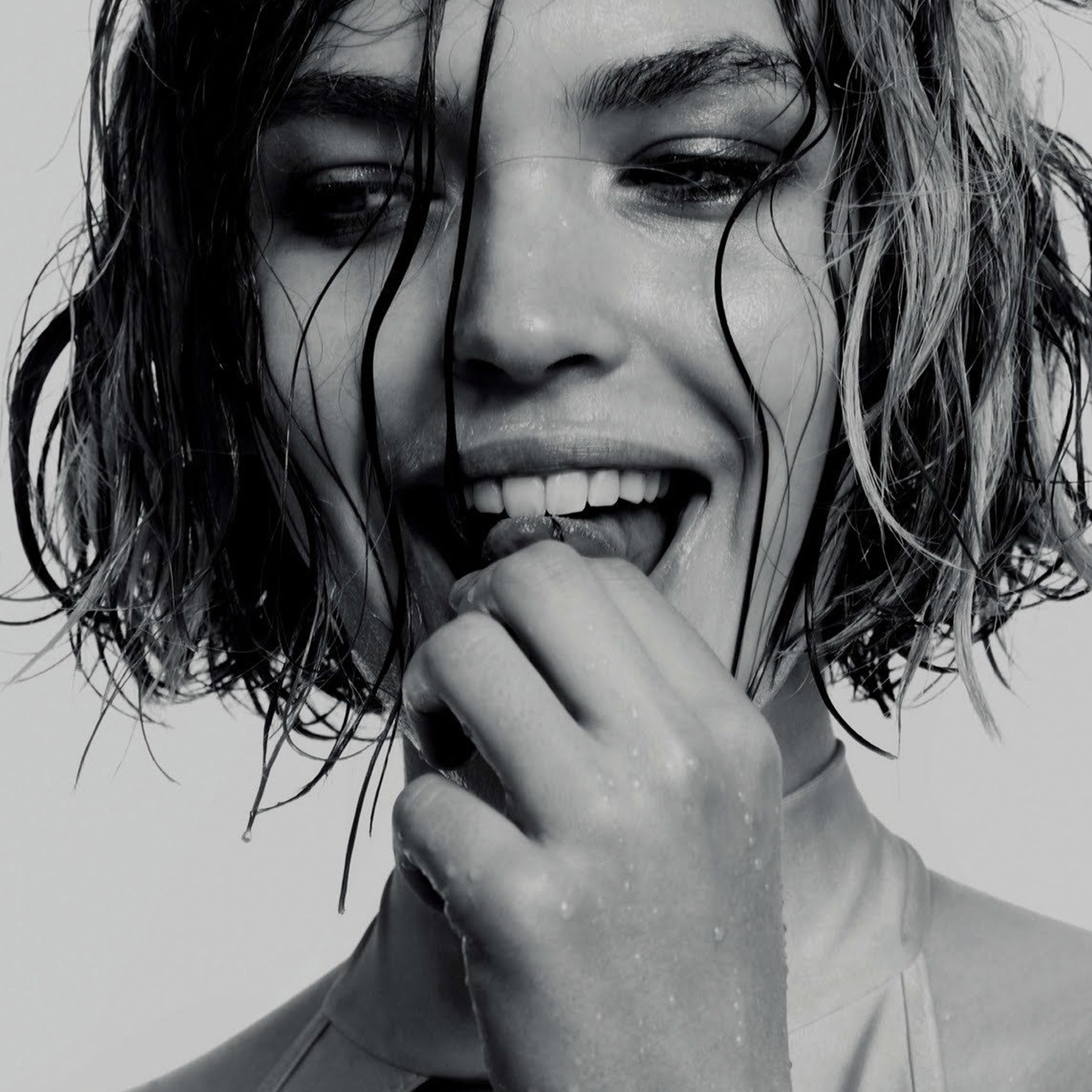 Close-up black-and-white photo of a woman with wet, curly hair, smiling with her eyes closed, biting her finger.