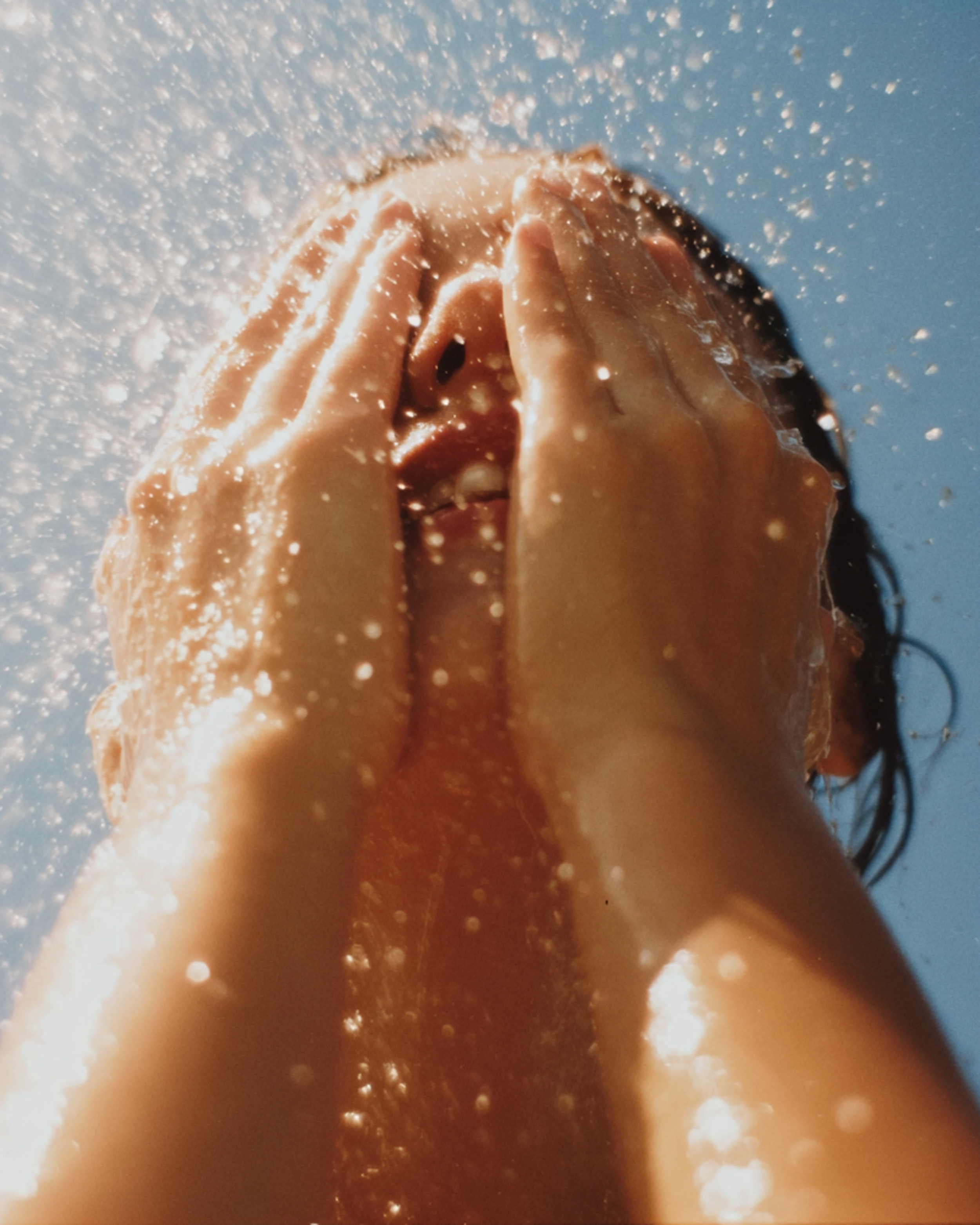 A person taking a photo while holding their nose, with a blue sky and sunlight in the background, as water spray or mist surrounds them.