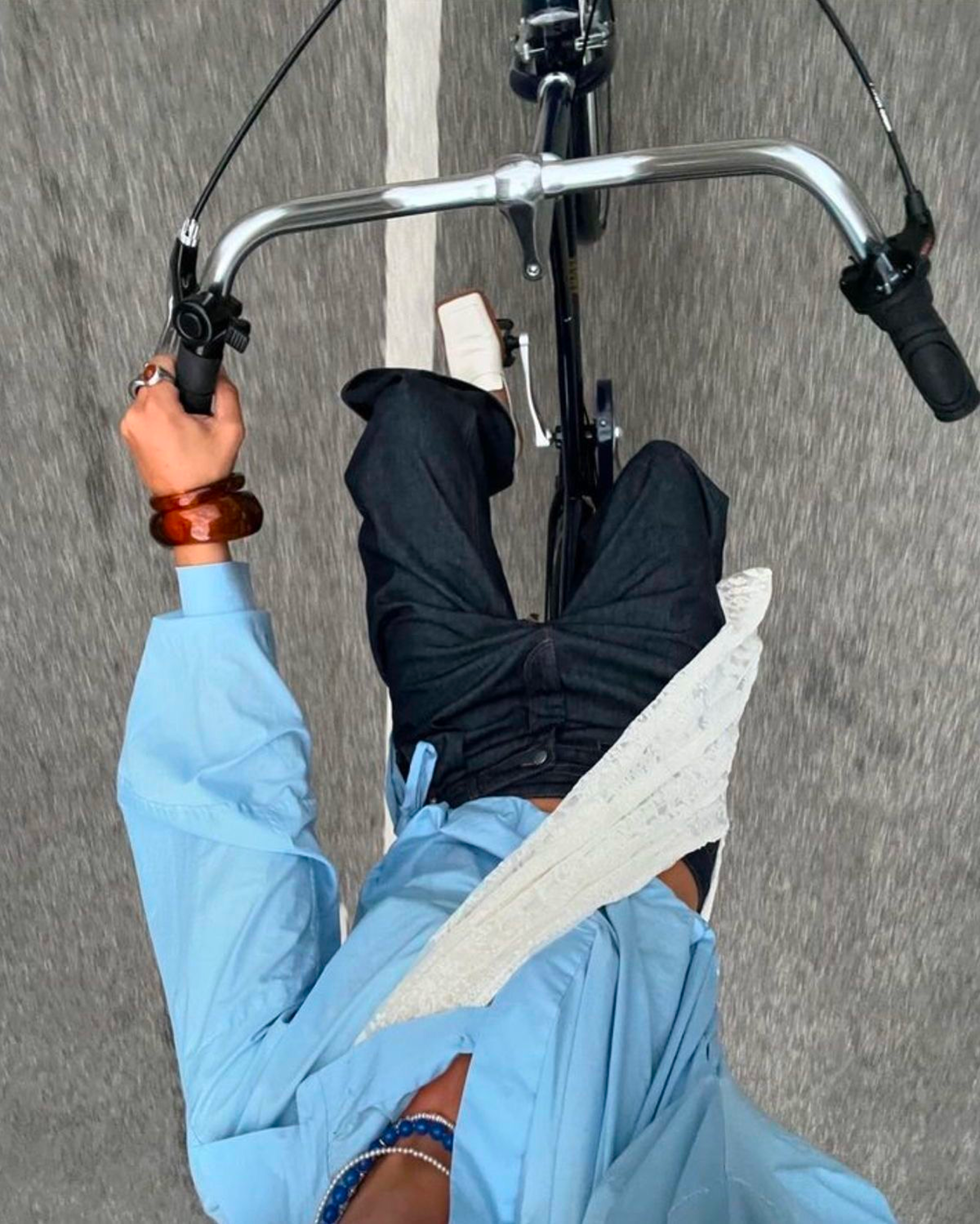 A person in a yellow top, blue bracelet, and white pants walking on a gray road with a white dashed line, viewed from above. The person is holding handlebars of a scooter or bicycle and has a bandage on their knee.
