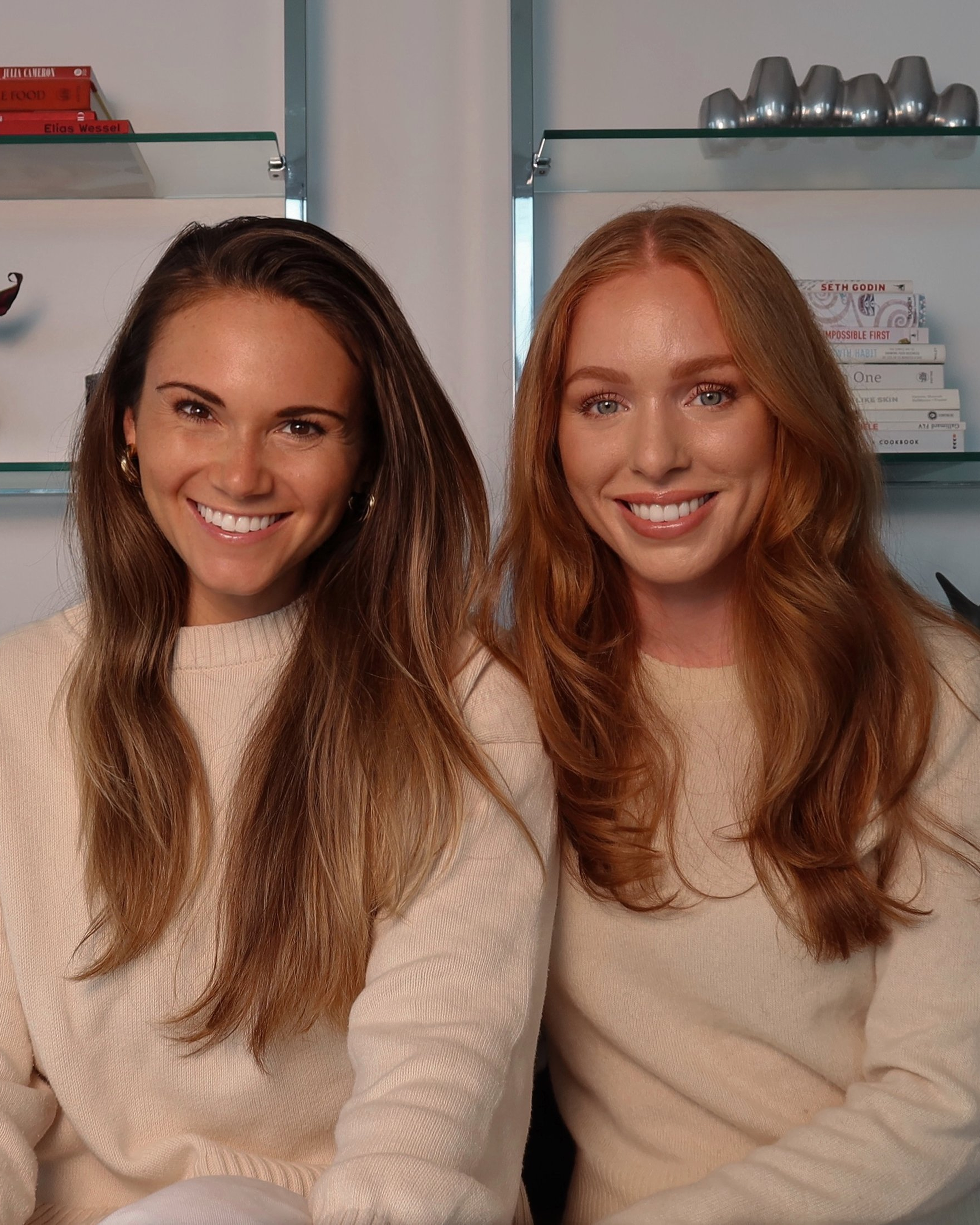 Close-up of smiling women with long hair, wearing beige sweaters, in an indoor setting with shelves and books in the background.