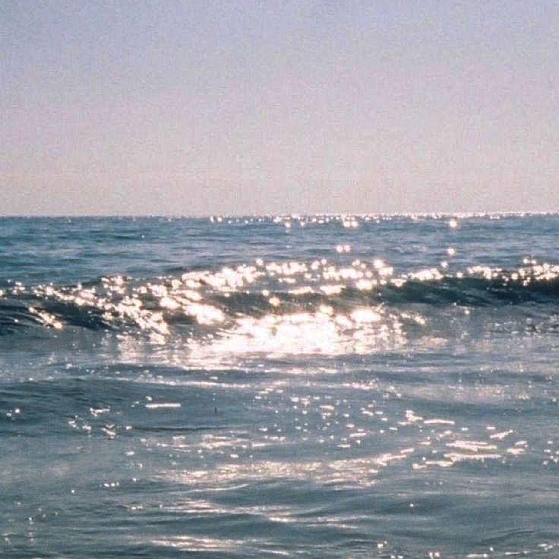 A wide view of the ocean with small waves and the sun reflecting off the water, with a clear sky in the background.