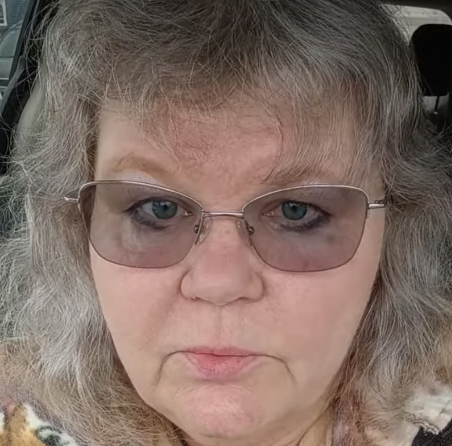 Close-up of a woman with gray hair and wearing sunglasses, with a neutral facial expression.