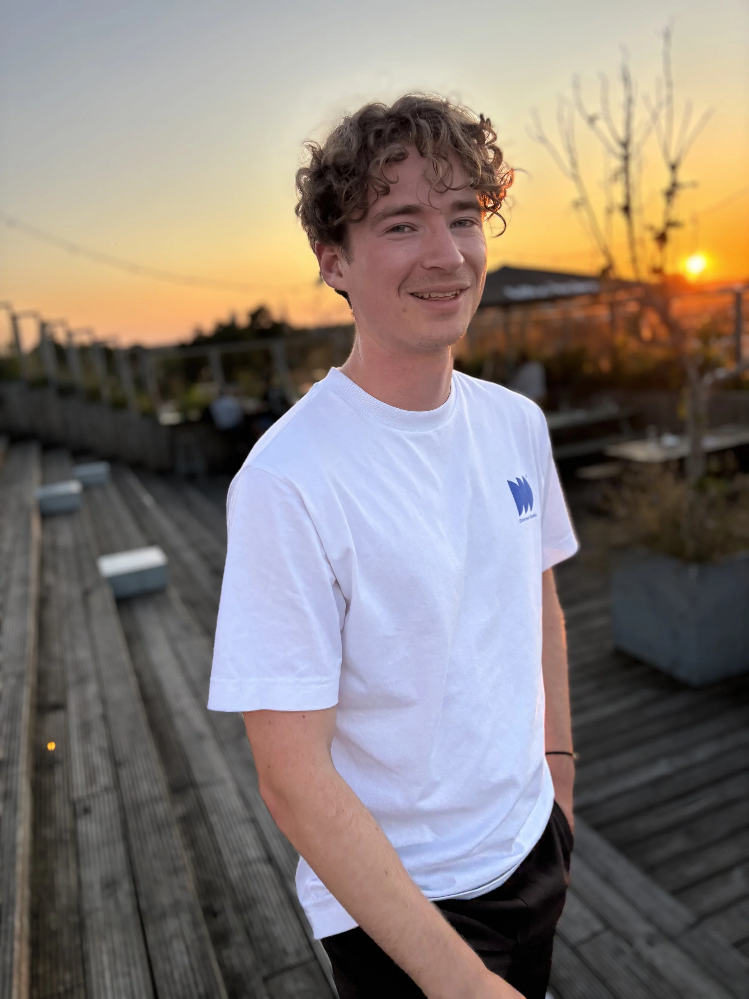 Portret van een jonge man met krullend haar, lachend, buiten tijdens zonsondergang op een houten terras.