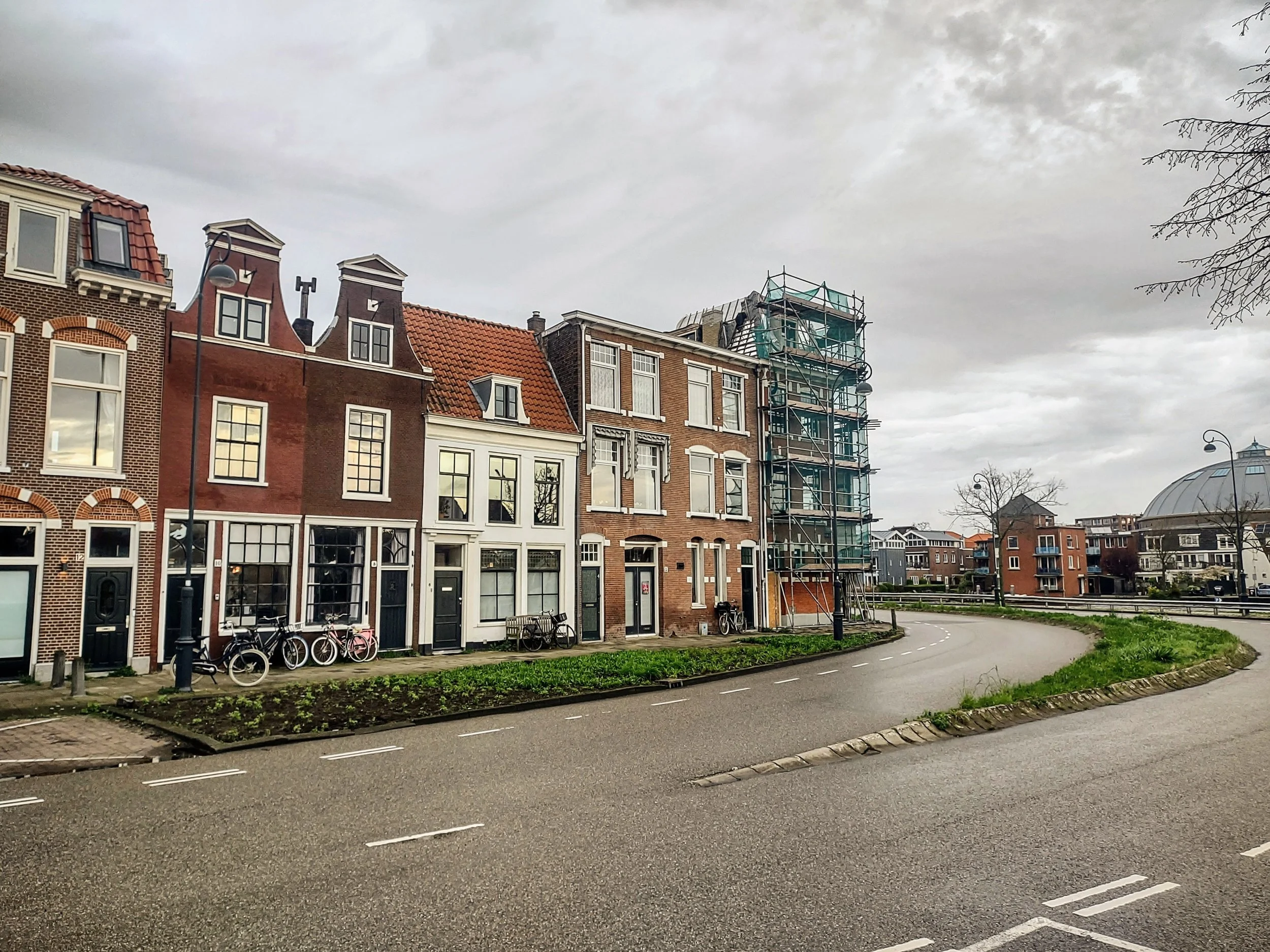 Rijen oude Nederlandse herenhuizen met verschillende gevels en en rooftypen, een steiger voor een gebouw onder renovatie, langs een straat met fiets- en parkeerplaatsen onder een bewolkte hemel.