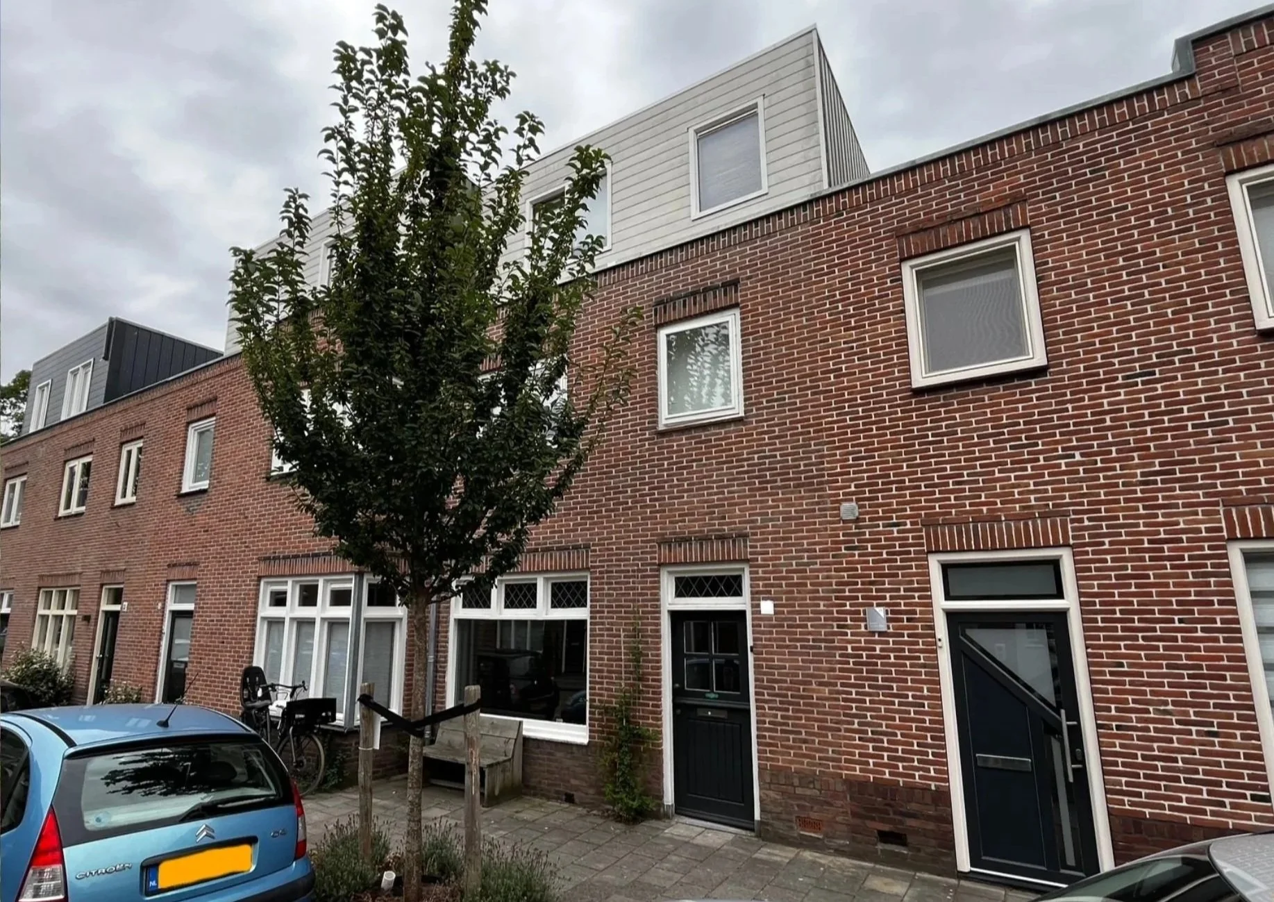 Een rij bakstenen gevelwoningen met ramen en deuren, een blauwe hemel en een straat met personeelsauto's.