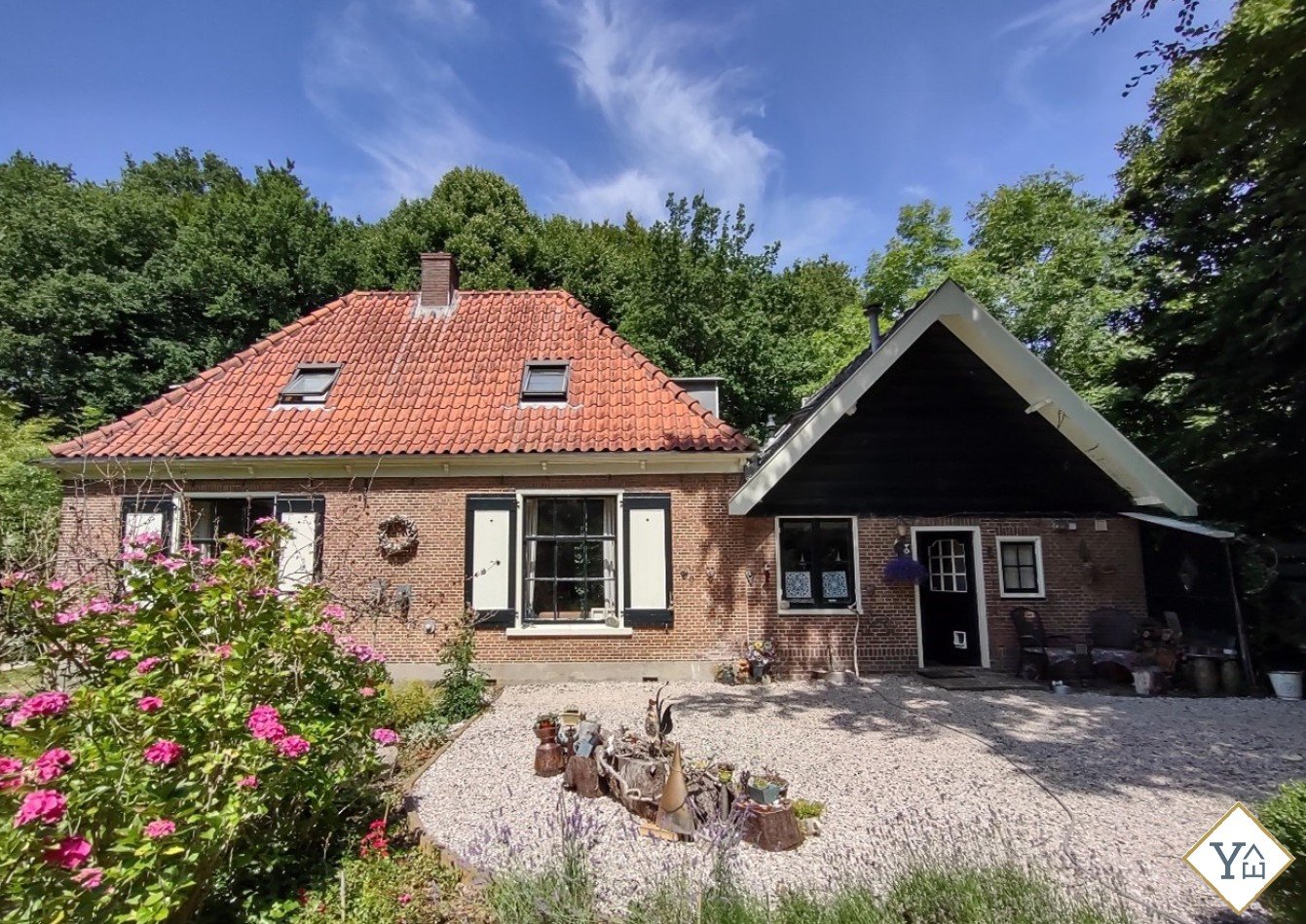 Een traditionele Nederlandse woning met een rode dakpannen en een houten aanbouw aan de rechterkant onder een blauwe hemel met wolken. Er staan bloemen en planten voor het huis.