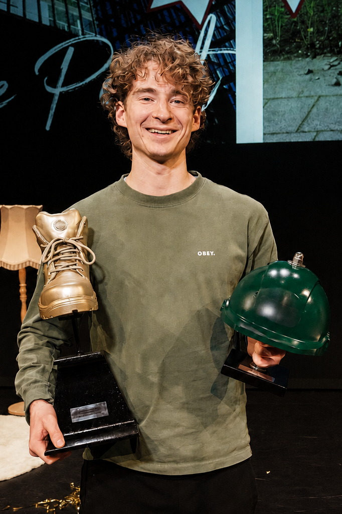 Jonge man met curly haar die een prijs ontvangt, vasthoudend een gouden laarzen-beeld en een groene cloche-helm op standaards, op een podium met een achtergrond met grafische elementen.