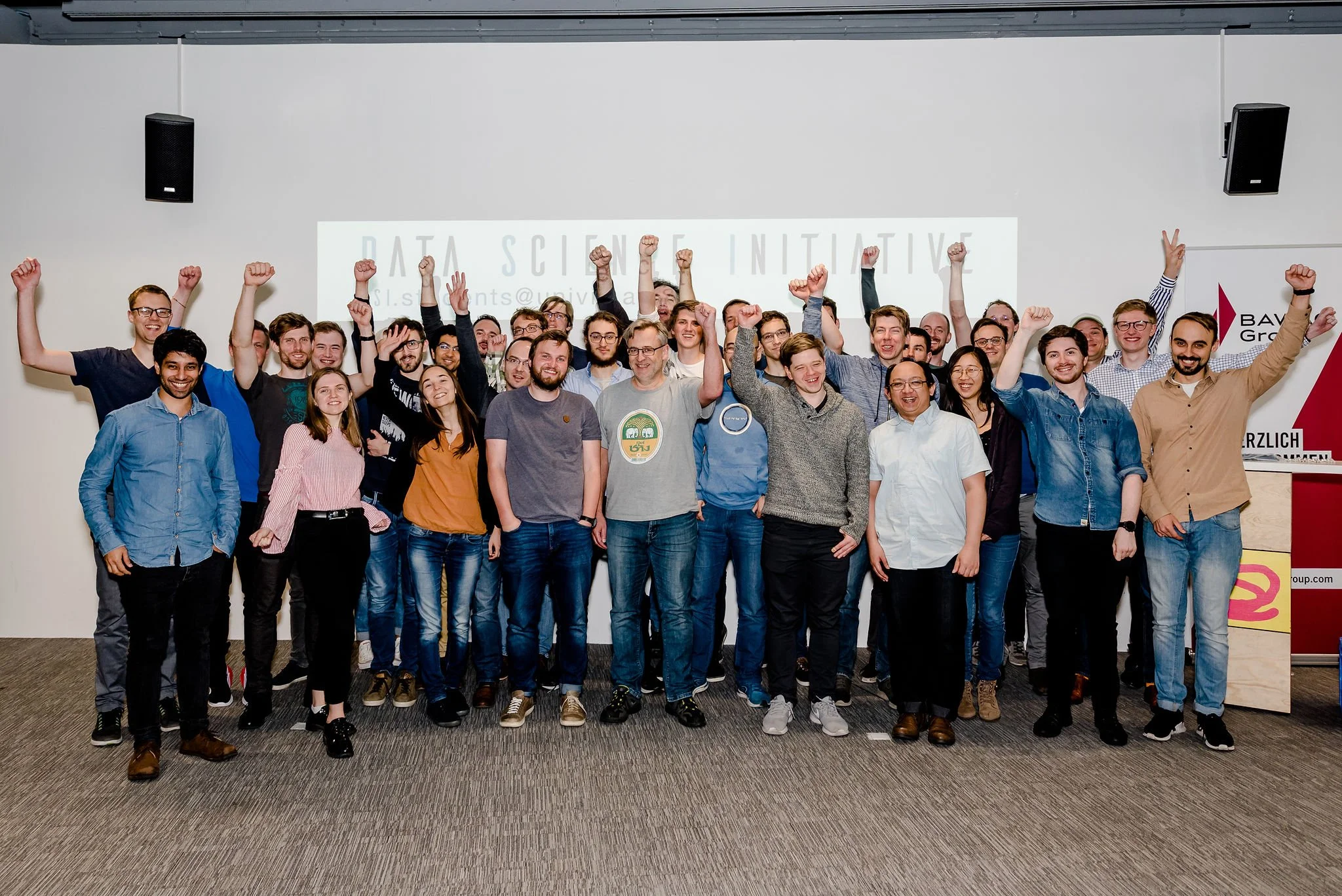 Gruppe junger Menschen auf einer Konferenzbühne, die jubeln und die Arme in die Luft strecken, vor einem weißen Bildschirm mit der Aufschrift 'Data Science Initiative'.