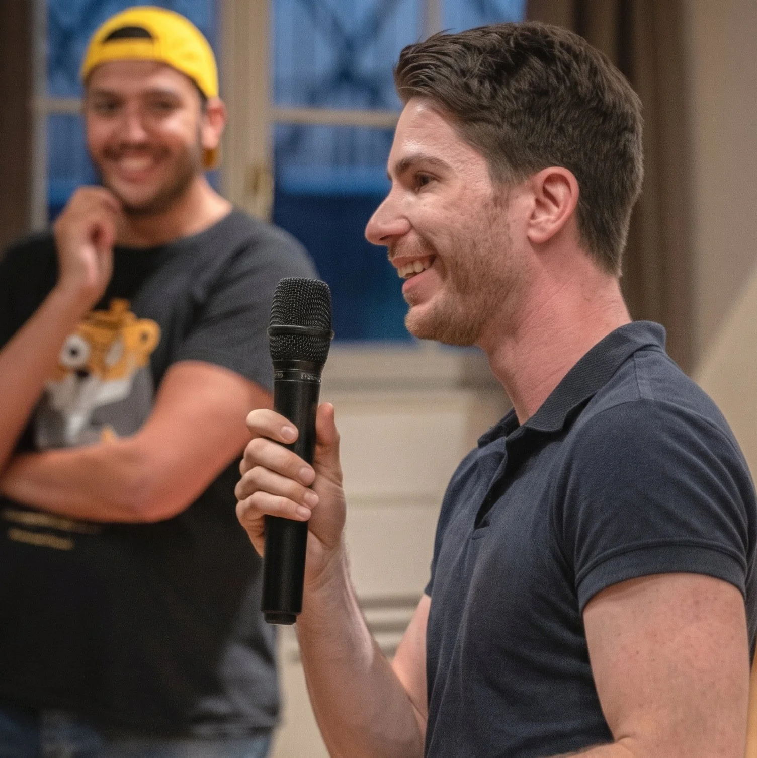 Ein Mann mit dunklem Haar und Bart hält ein Mikrofon, während er spricht. Im Hintergrund steht ein lachender junger Mann mit hellbraunem Haar, der eine schwarze T-Shirt trägt und eine gelbe Kappe trägt.