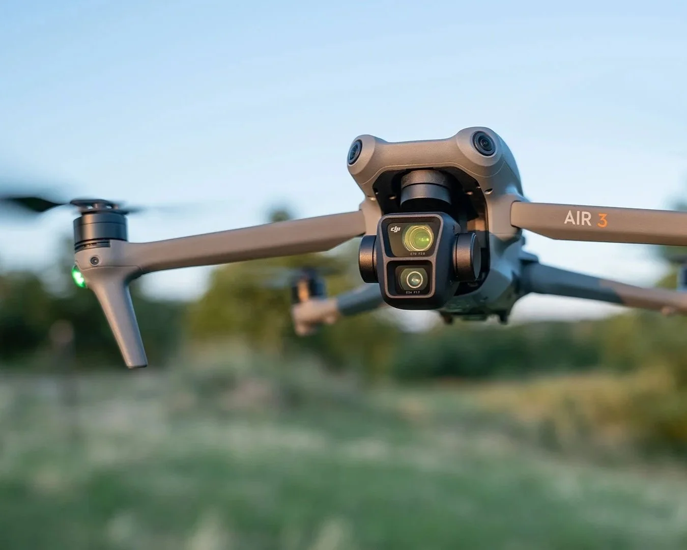 Close-up of a DJI Air 3 drone flying outdoors with a background of trees and a clear sky, during daytime.