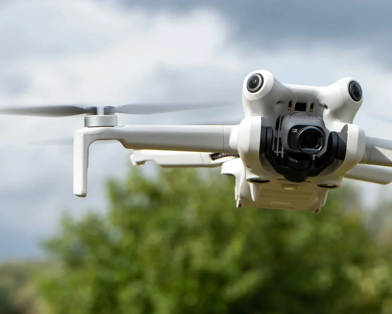 A white quadcopter drone with a mounted camera flying outdoors against cloudy sky and green foliage.
