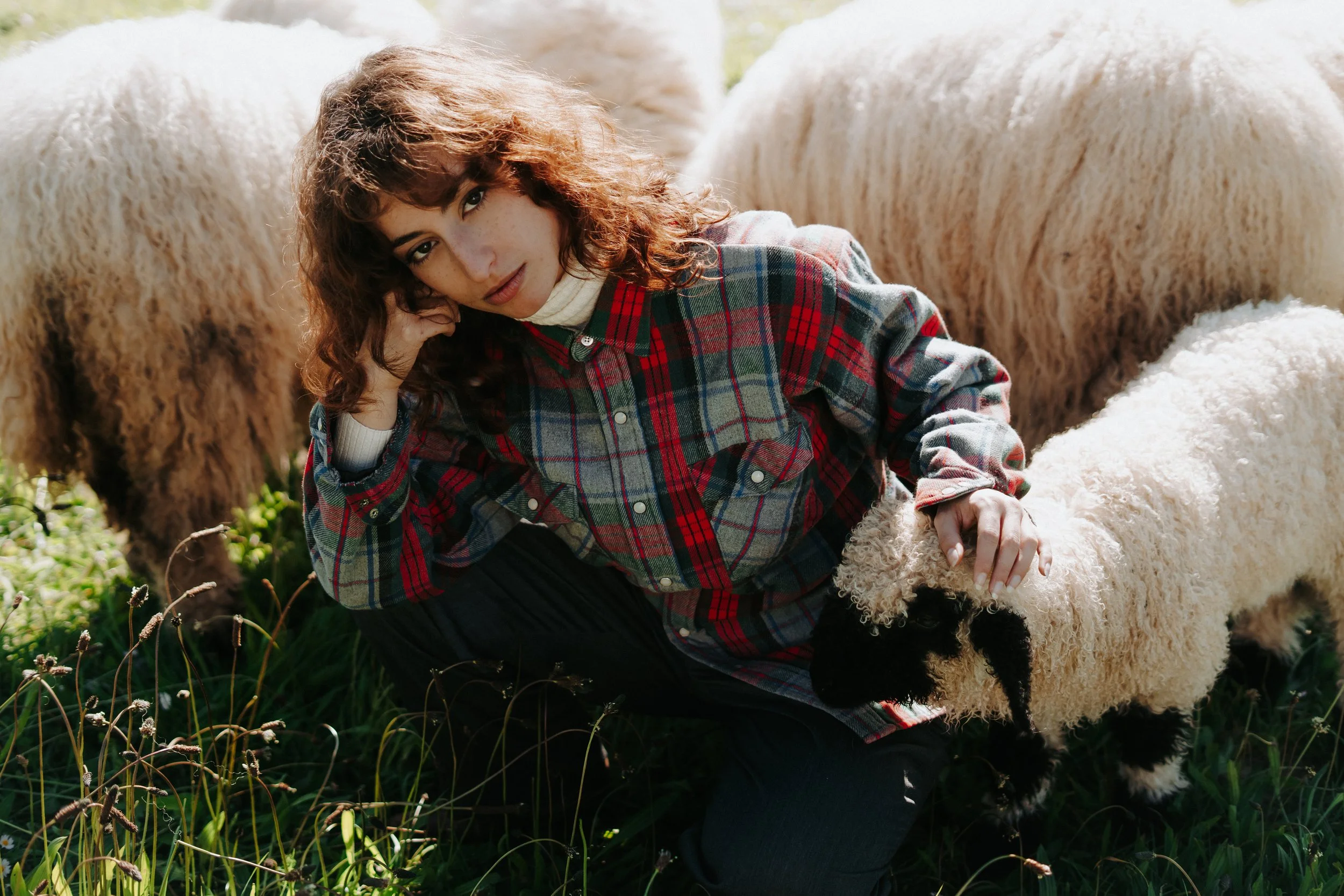 Une femme avec des cheveux bouclés, portant un manteau en tartan rouge et vert, caresse un agneau blanc dans un champ vert.