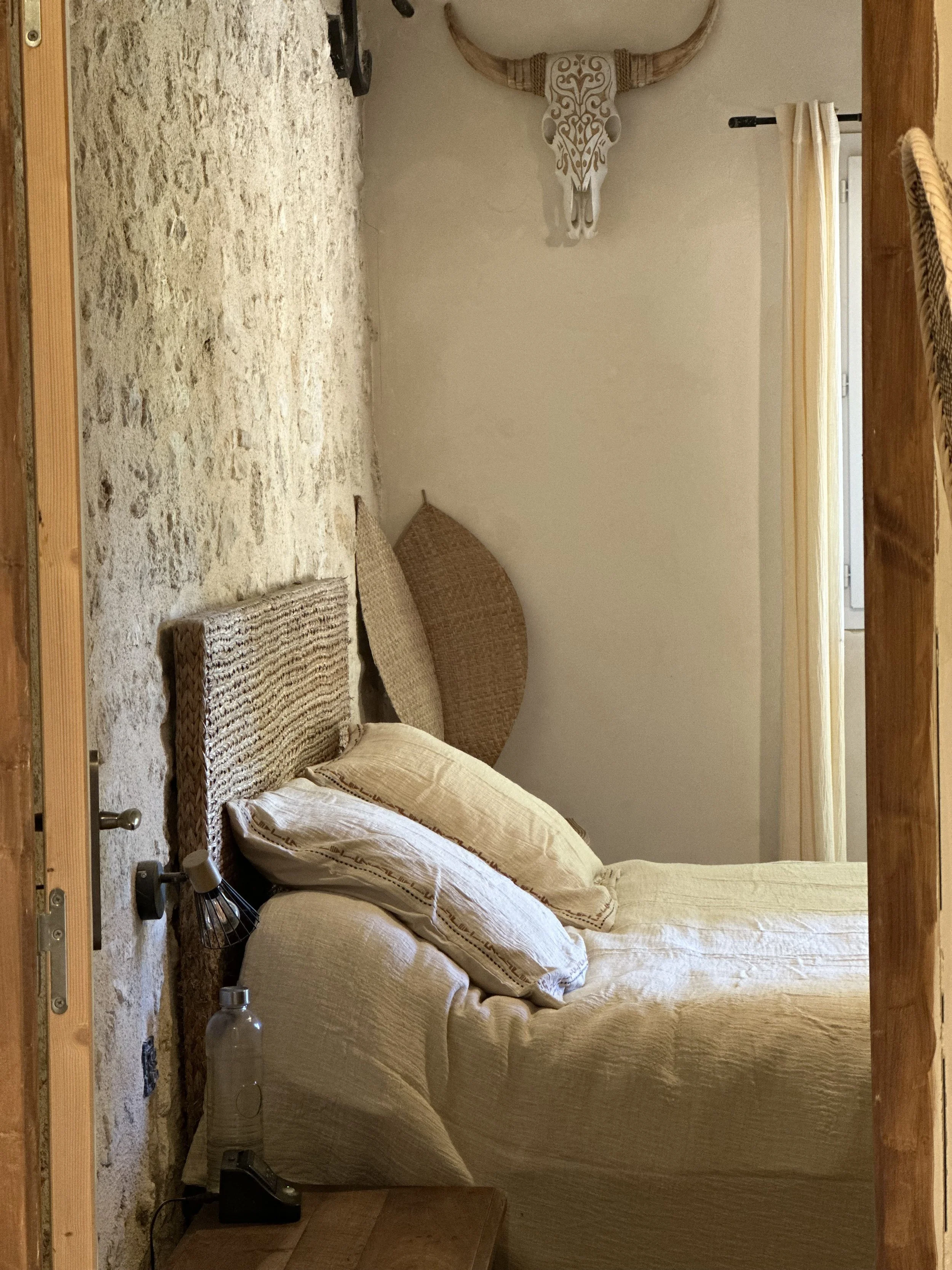 Une pièce avec un mur de pierre, un canapé beige avec des coussins, une tête de taureau en décoration sur le mur, une table en bois, une lampe de bureau, une bouteille d'eau en plastique sur le sol et une fenêtre avec rideau beige.
