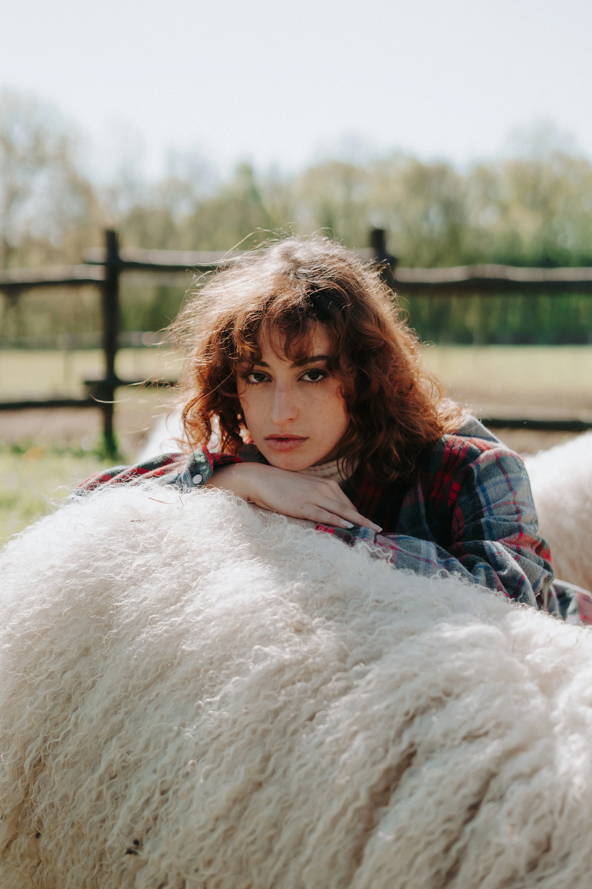 Une jeune femme avec des cheveux bruns bouclés, portant un chemisier à carreaux rouge et bleu, repose sur une peau de mouton blanche à l'extérieur dans un champ, avec un arbre en arrière-plan et un ciel clair.