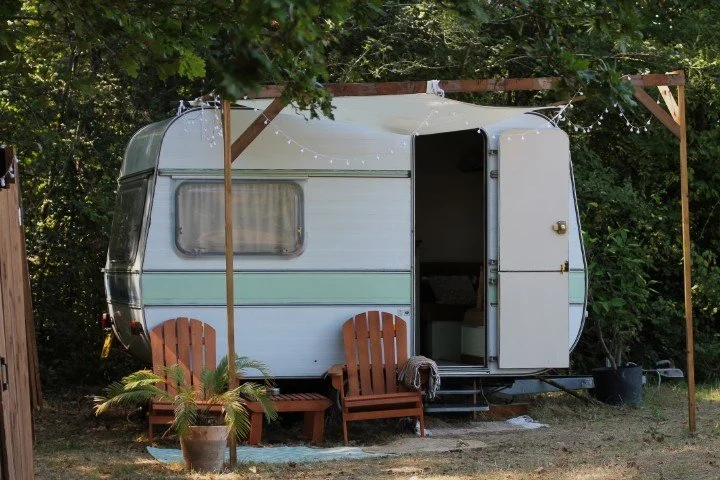 Une caravane blanche sous un abri en bois avec deux chaises en bois devant, entourée d'arbres et de plantes, avec des guirlandes lumineuses suspendues.