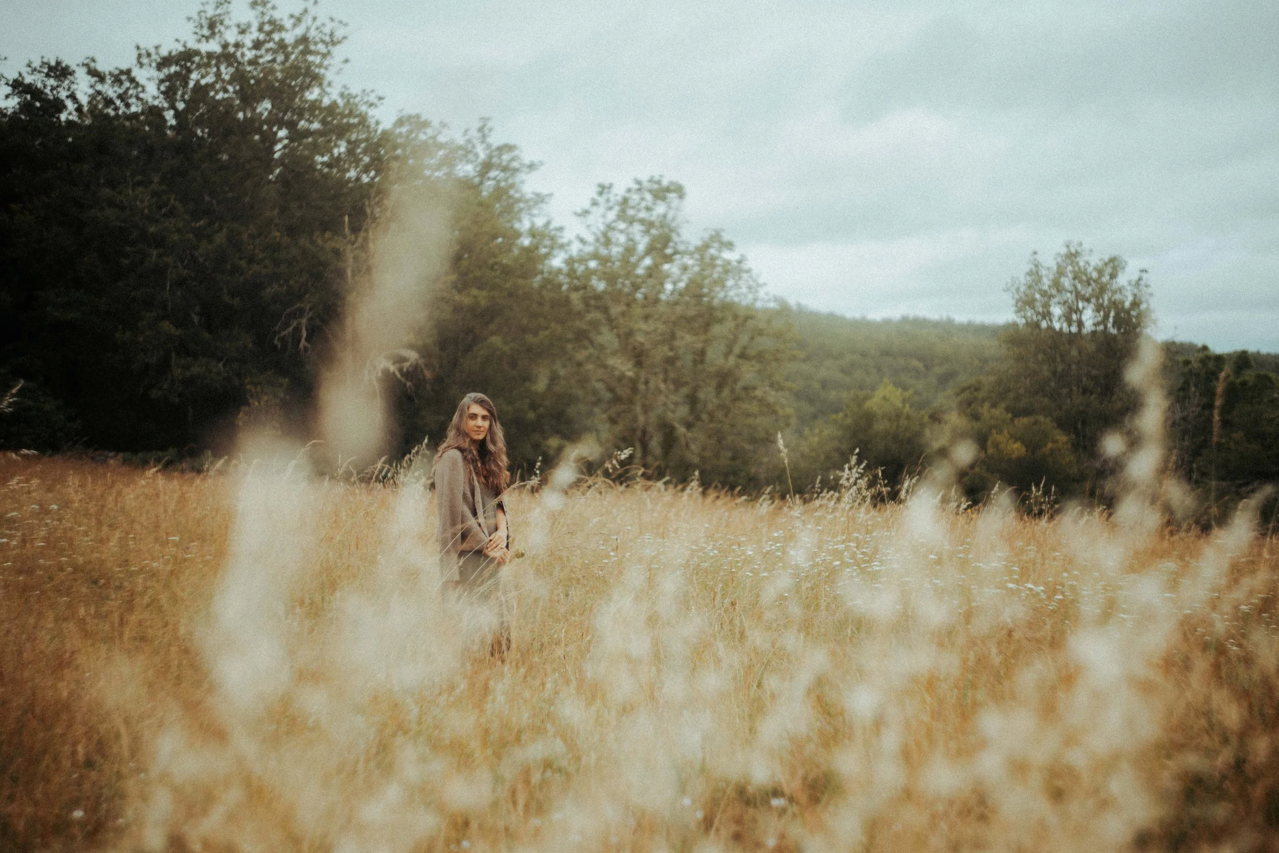 Une femme dans un champ d'herbes hautes, avec des arbres en arrière-plan et un ciel nuageux.