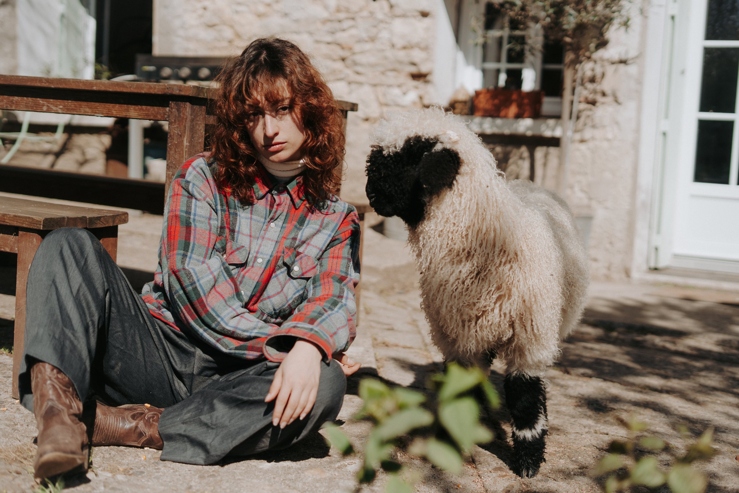 Une femme aux cheveux bouclés roux, habillée d'une chemise à carreaux, assise sur le sol à côté d'un chien de berger australien avec un pelage épais et bouclé, devant une maison en pierre. Elle regarde vers l'objectif dans un cadre extérieur ensoleil