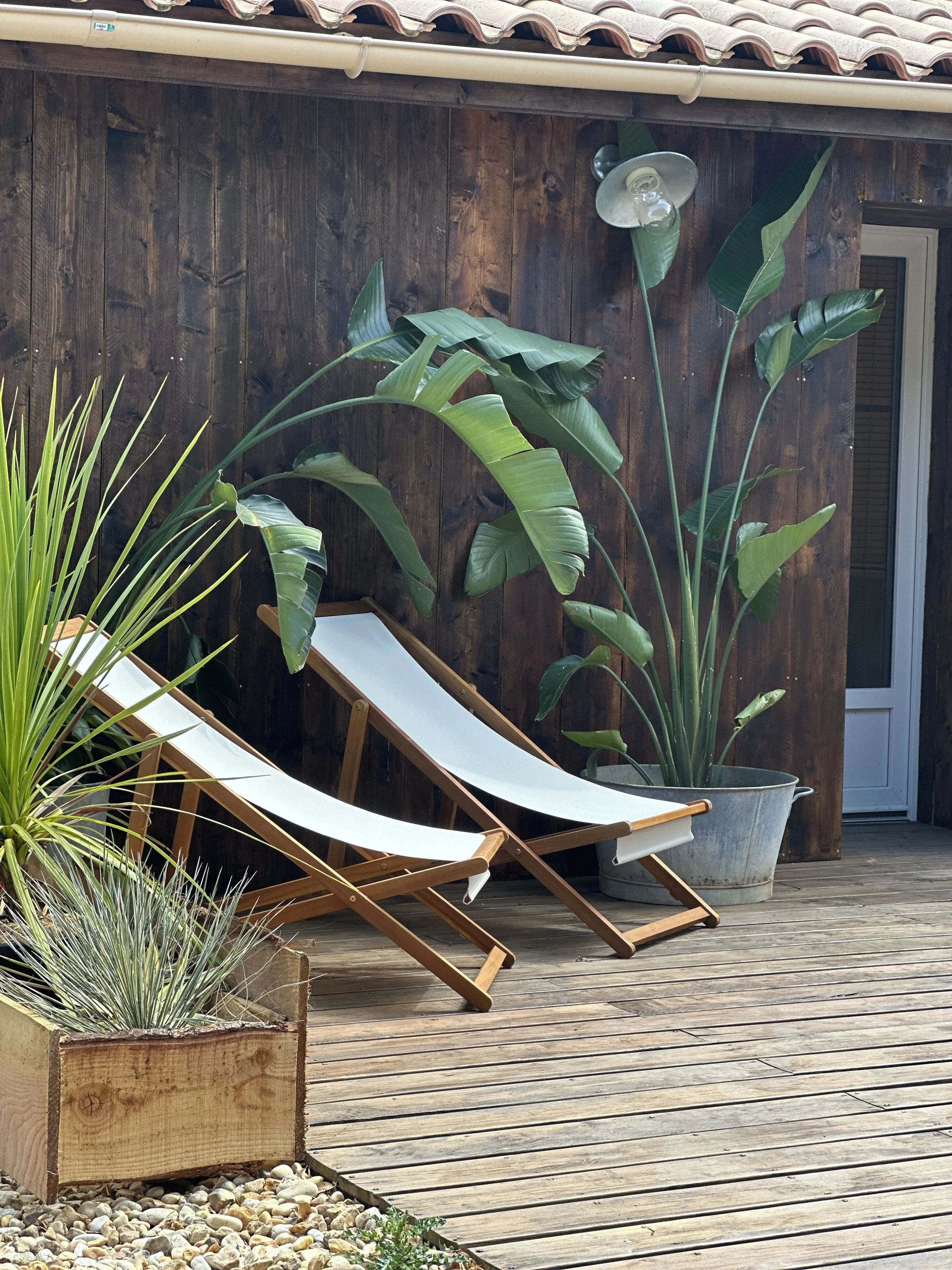 Une terrasse en bois avec deux chaises longues en bois et toile blanche, un grand pot avec une plante verte, un mur en bois foncé et une fenêtre en arrière-plan. Plus à gauche, une jardinière en bois contenant diverses plantes, dont une graminée. Un éclairage de jardin est fixé sur le mur en bois.