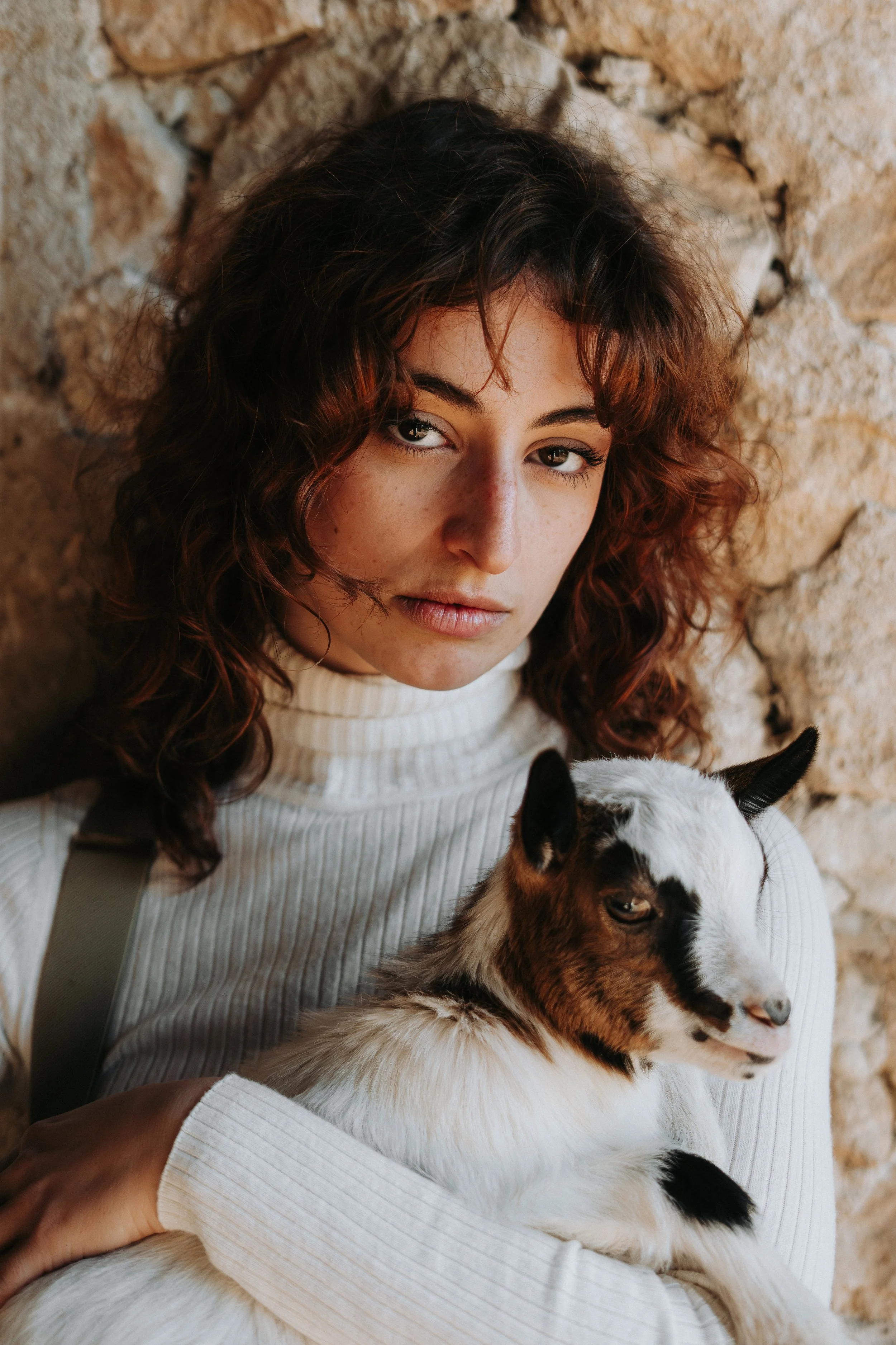 Une jeune femme aux cheveux bouclés et bruns, portant un pull beige, tient un bébé chèvre blanc avec des taches brunes, contre un mur en pierre.