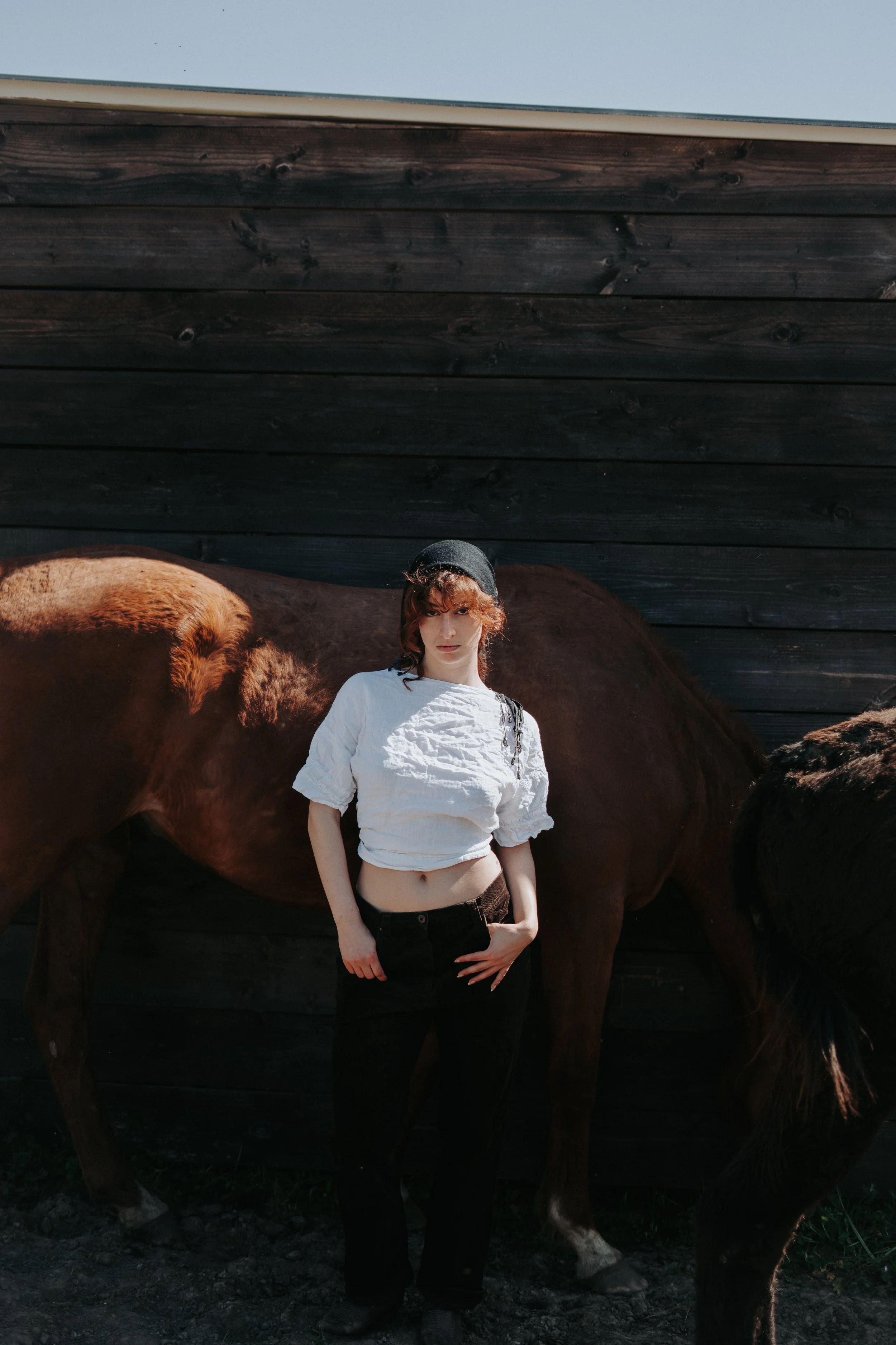 Une femme devant une clôture en bois, avec un cheval marron à ses côtés, portant un pull blanc, un béret noir et un pantalon noir.