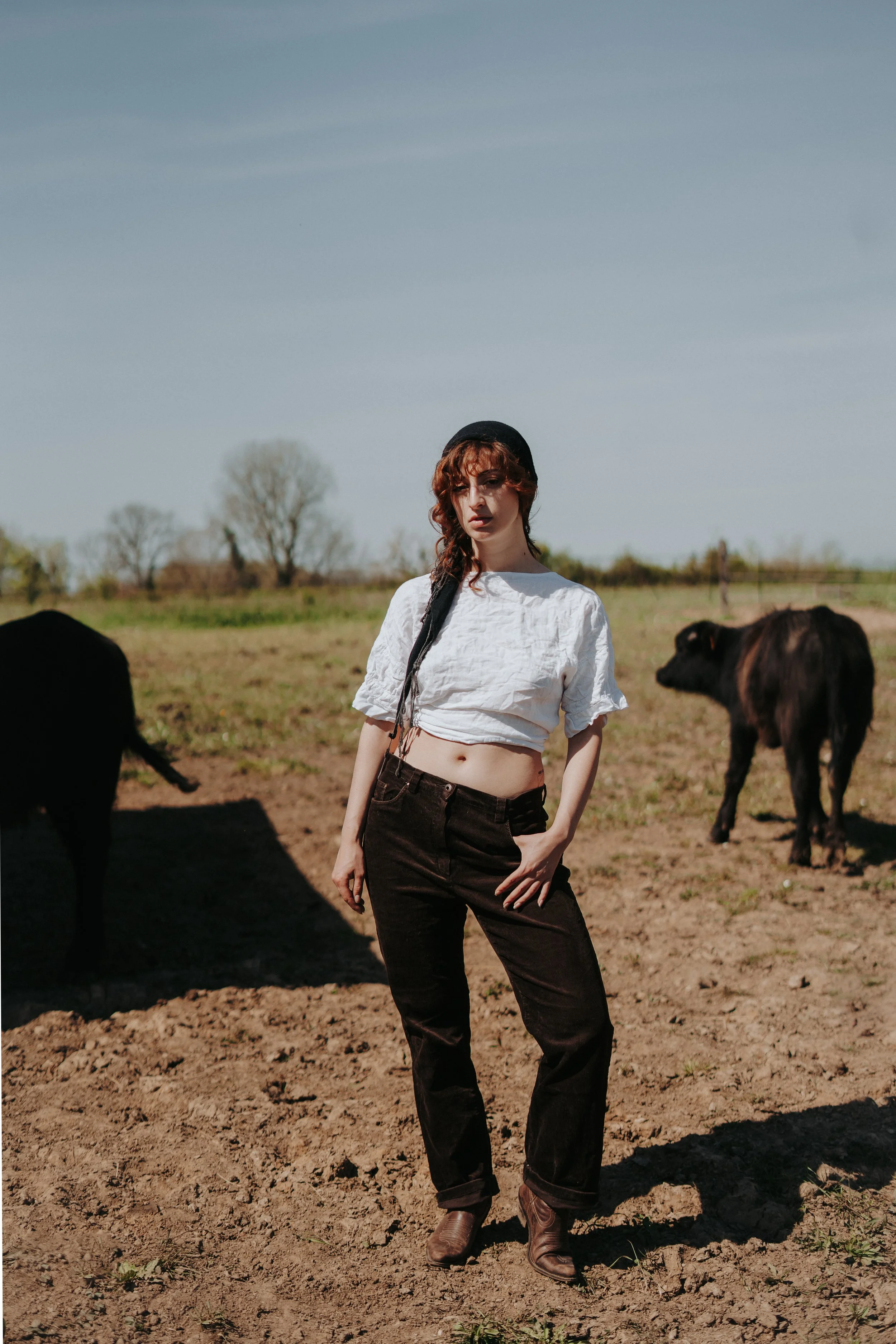Jeune femme portant un chapeau noir et un crop top blanc, se tenant dans un champ avec des vaches, sous un ciel dégagé.