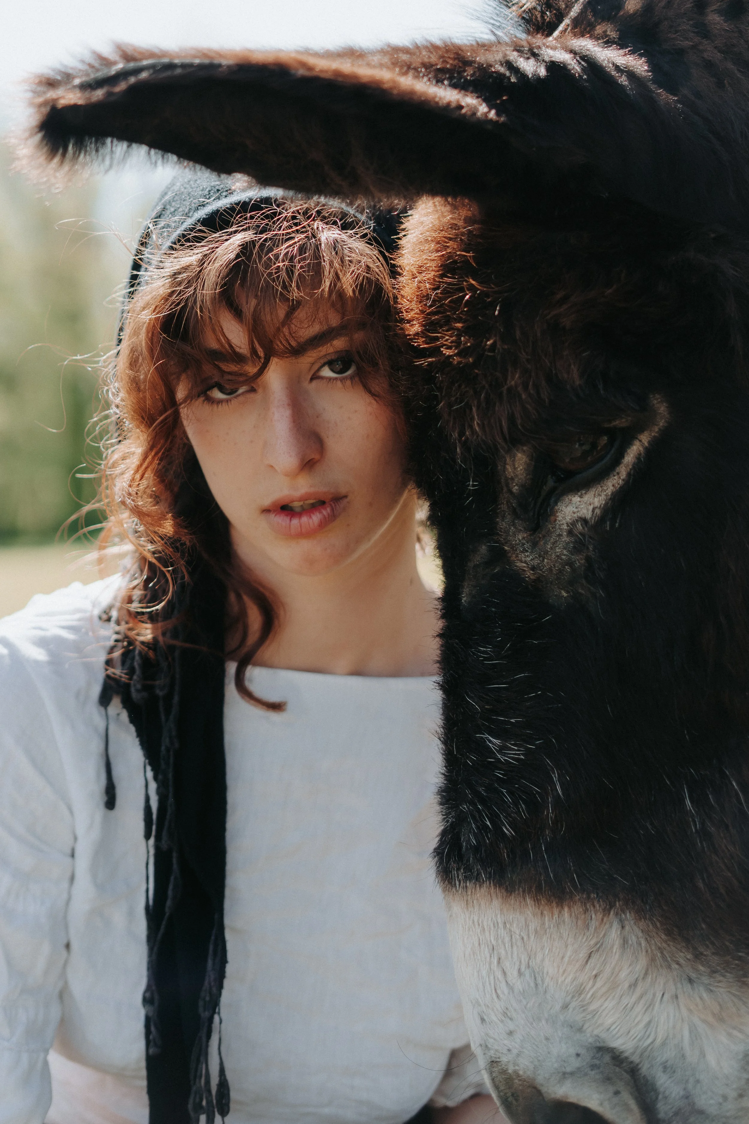 Une jeune femme avec des cheveux bouclés et une expression neutre, portant un haut blanc, prend une photo avec un cheval noir et blanc dans un environnement extérieur.