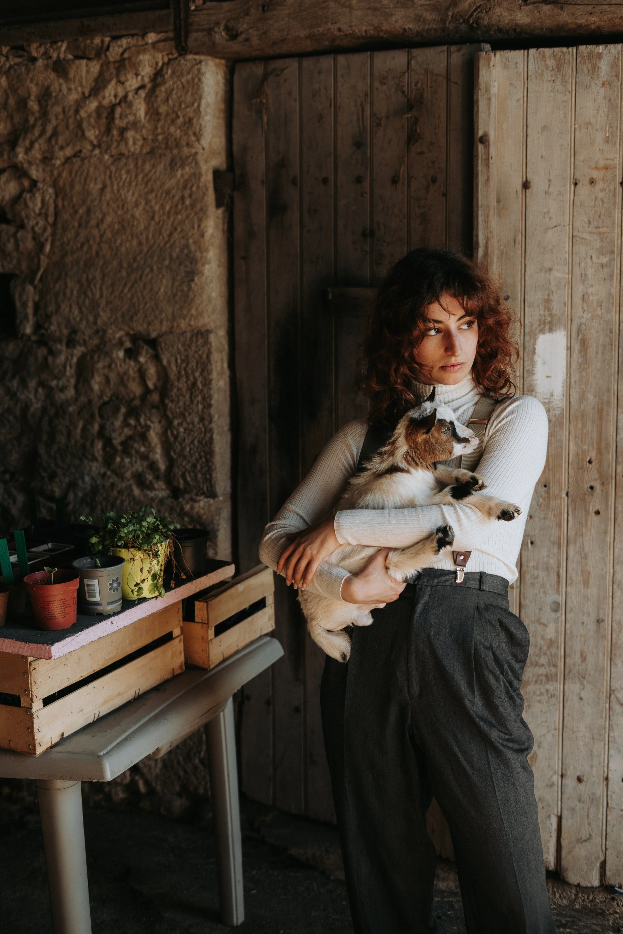 Une femme aux cheveux bouclés roux tient un chiot border collie dans ses bras, dans un intérieur rustique avec des murs en pierre et en bois, et une table avec des plantes en pots.