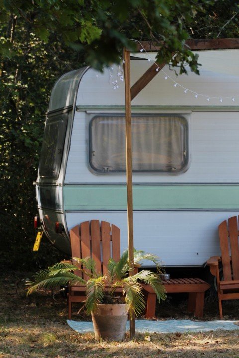 Une caravane blanche avec des rideaux en polyester derrière deux chaises en bois, une grande auge avec une plante verte, et un parasol en bois avec des guirlandes lumineuses dans un environnement extérieur verdoyant.