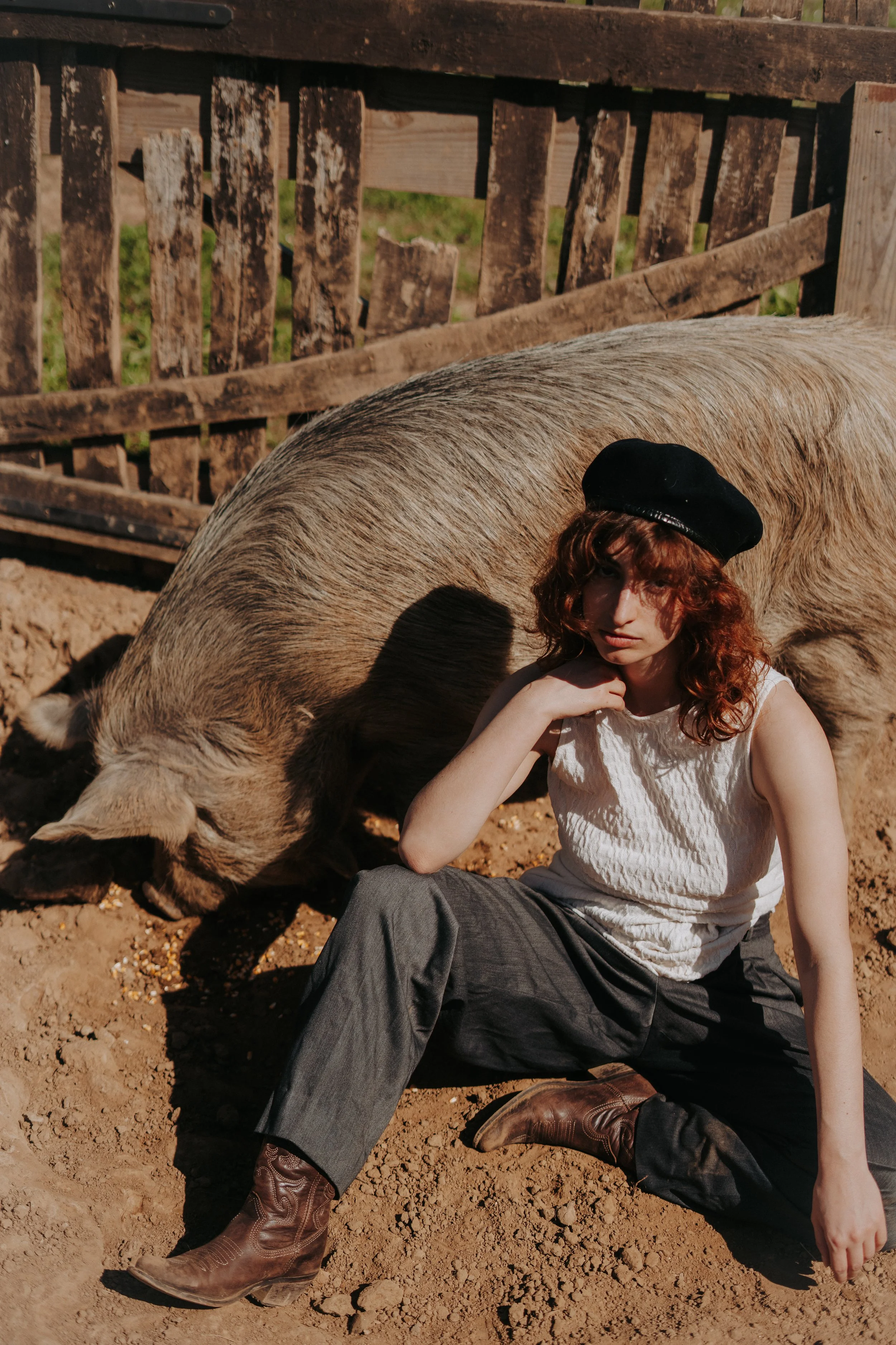 Une femme avec des cheveux bouclés rouges portant un béret noir, une chemise blanche à manches courtes et un pantalon gris foncé, assise dans une ferme avec un cheval gris tacheté, derrière une clôture en bois.