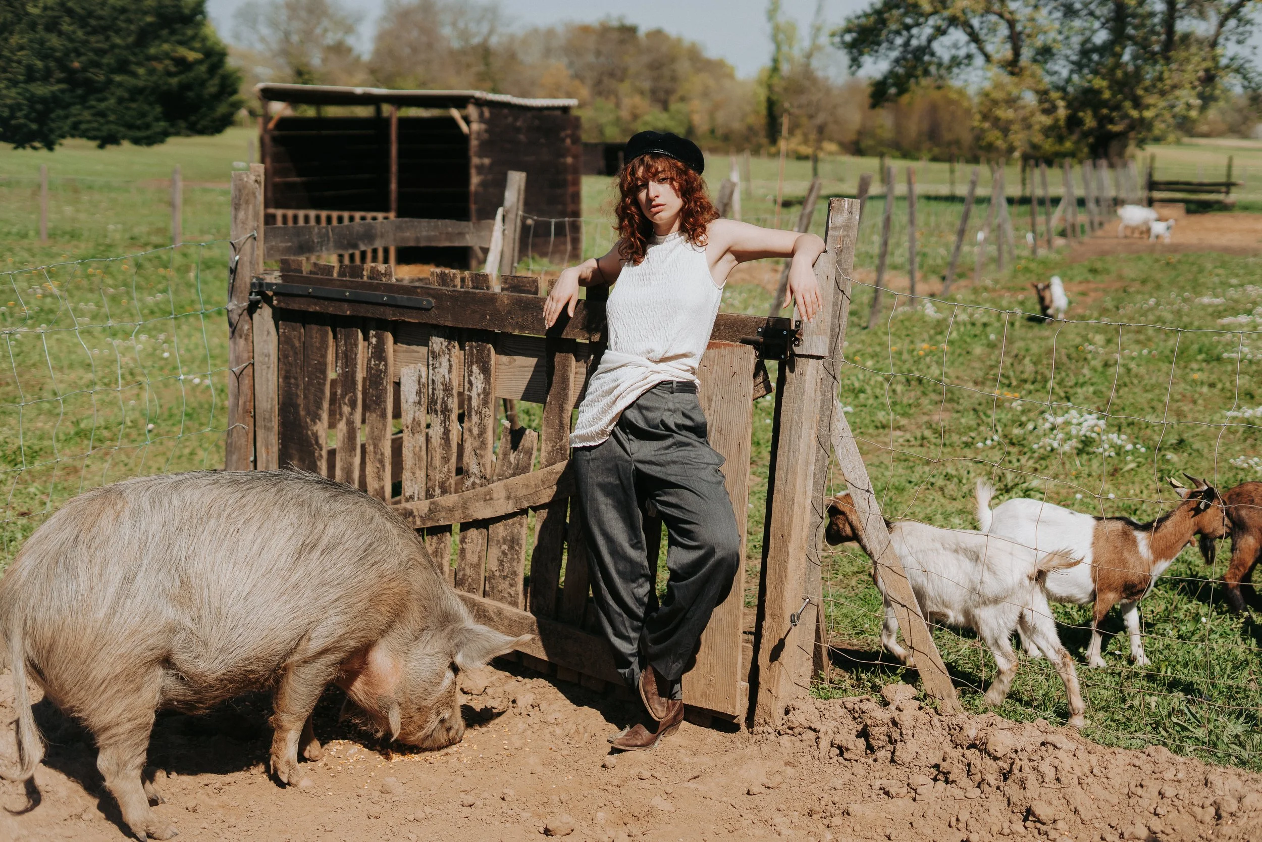 Une femme avec des cheveux bouclés, portant un chapeau noir, une chemise blanche et un pantalon gris, posant dans une ferme avec des chèvres et des cochons en arrière-plan, entourée d'une clôture en bois.