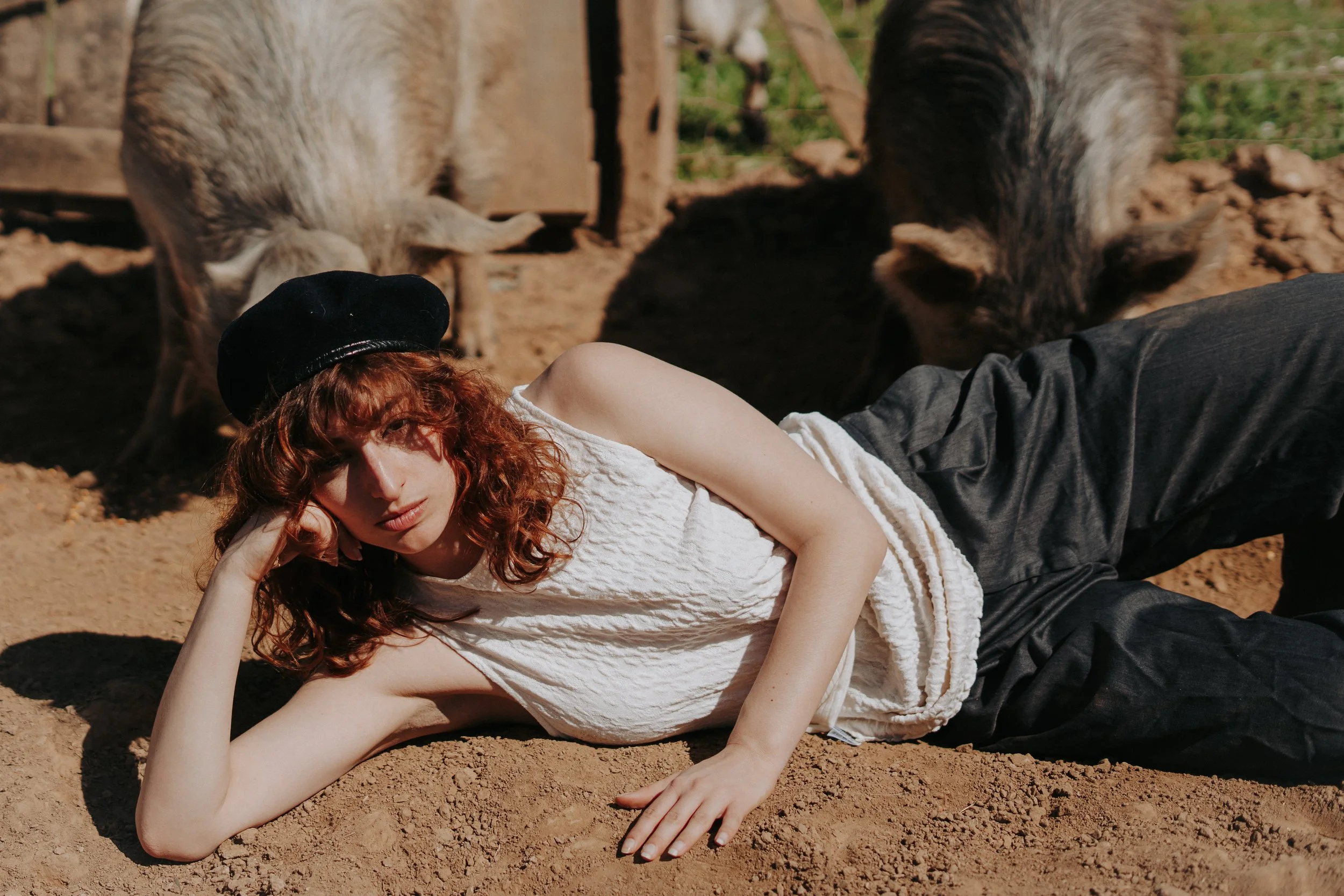 Jeune femme à cheveux roux, portant un beret noir, allongée sur la terre, entourée de porcs dans un enclos en plein air.