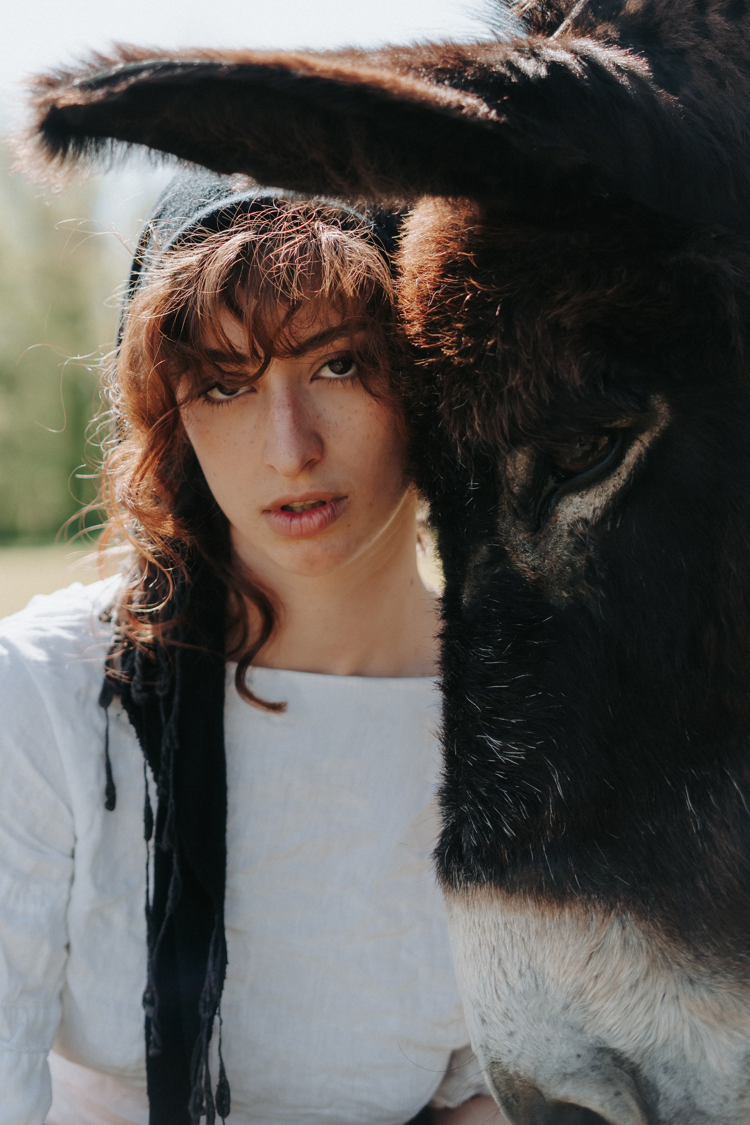 Une femme avec des cheveux bouclés et une chemise blanche pose à côté d'un cheval noir et blanc dont la tête est proche de la sienne.