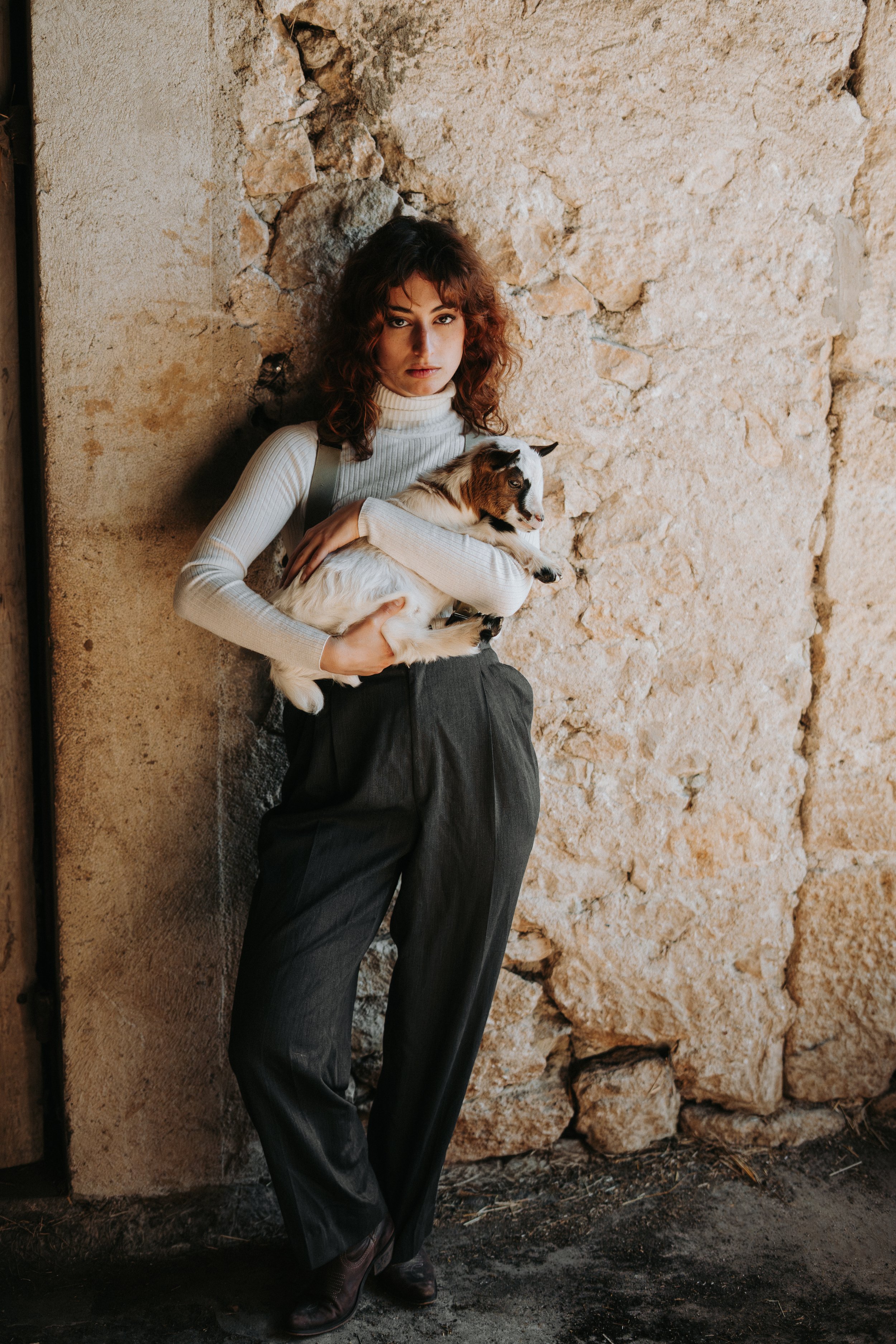 Une femme aux cheveux bouclés bruns portant un pull blanc et une jupe noire tient un petit chien blanc avec des taches brunes, devant un mur en pierre.