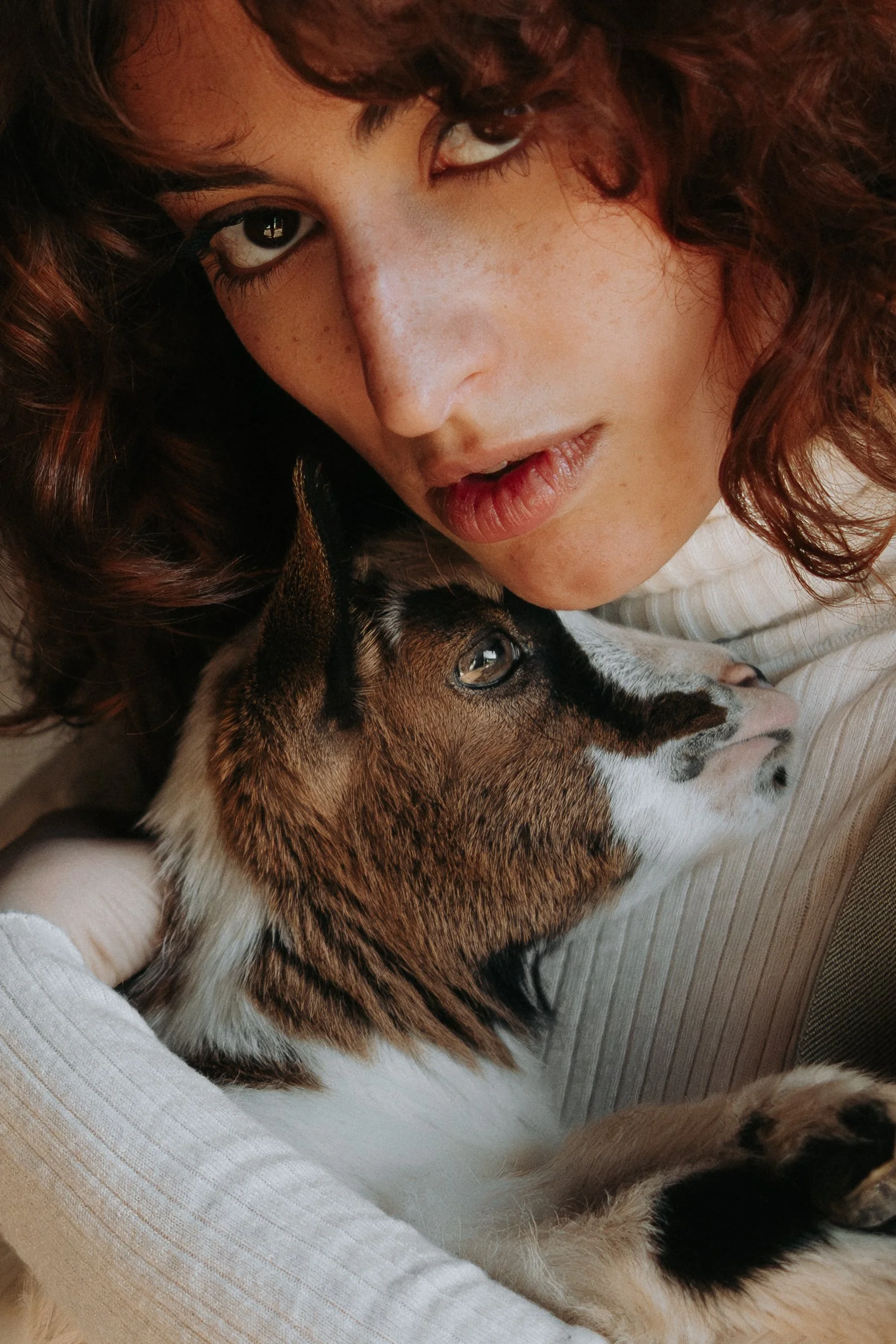 Une femme avec des cheveux bouclés bruns clairs et des taches de rousseur, tenant un chien à lacer, tous deux couchés sur un canapé beige.