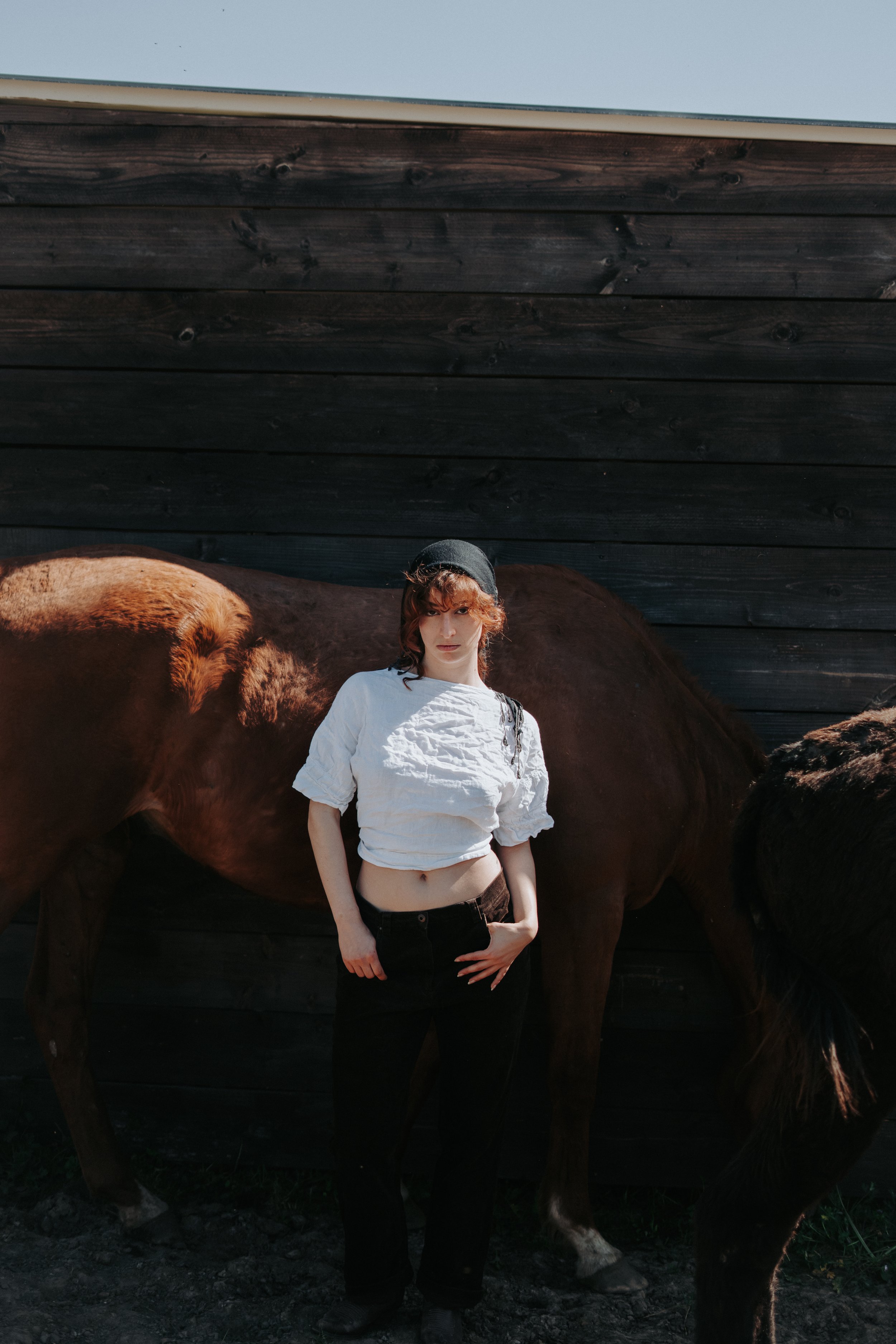 Une jeune femme avec un chapeau noir, une chemise blanche courte, et un pantalon noir, se tenant devant un cheval brun foncé avec un fond de plancher en bois noir.