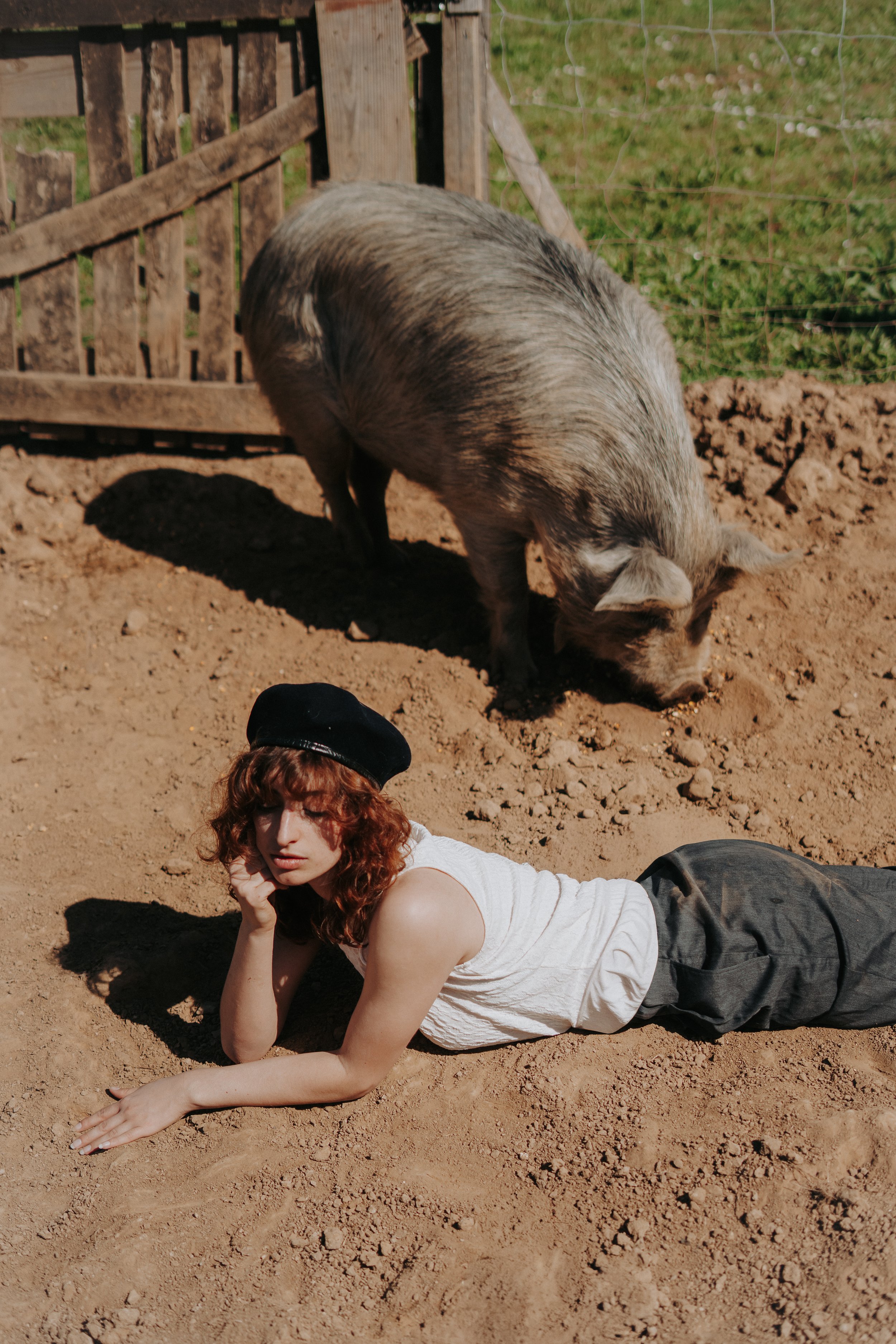Une femme aux cheveux bouclés roux, portant un haut blanc et un pantalon foncé, allongée sur le sol en terre, avec une main sous le visage, posée dans un champ en plein air. En arrière-plan, un cochon se trouve près d'une clôture en bois.