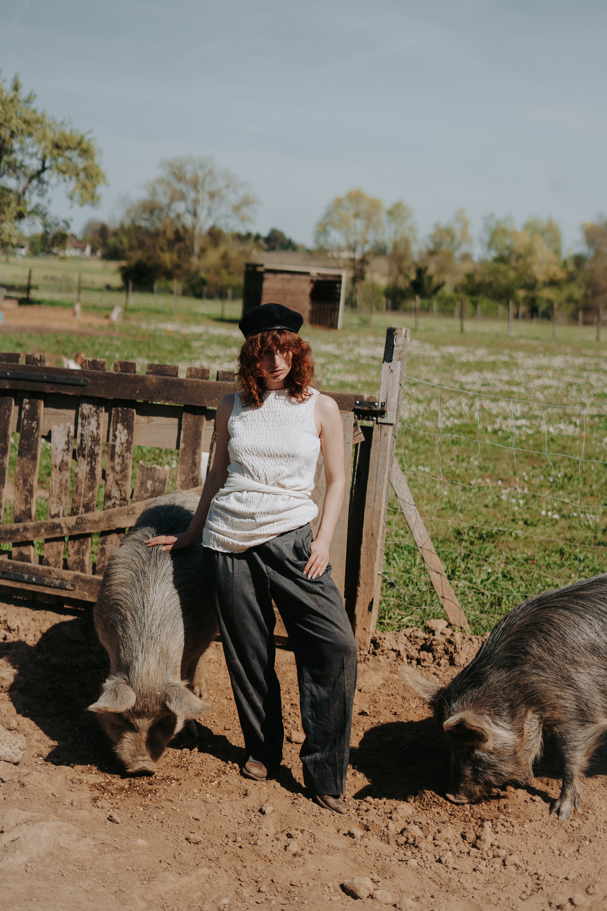 Une femme avec des cheveux bouclés portant un chapeau noir, une robe blanche et un pantalon noir, debout dans un enclos avec deux porcs à l'extérieur, dans un paysage rural avec des arbres et un ciel clair.