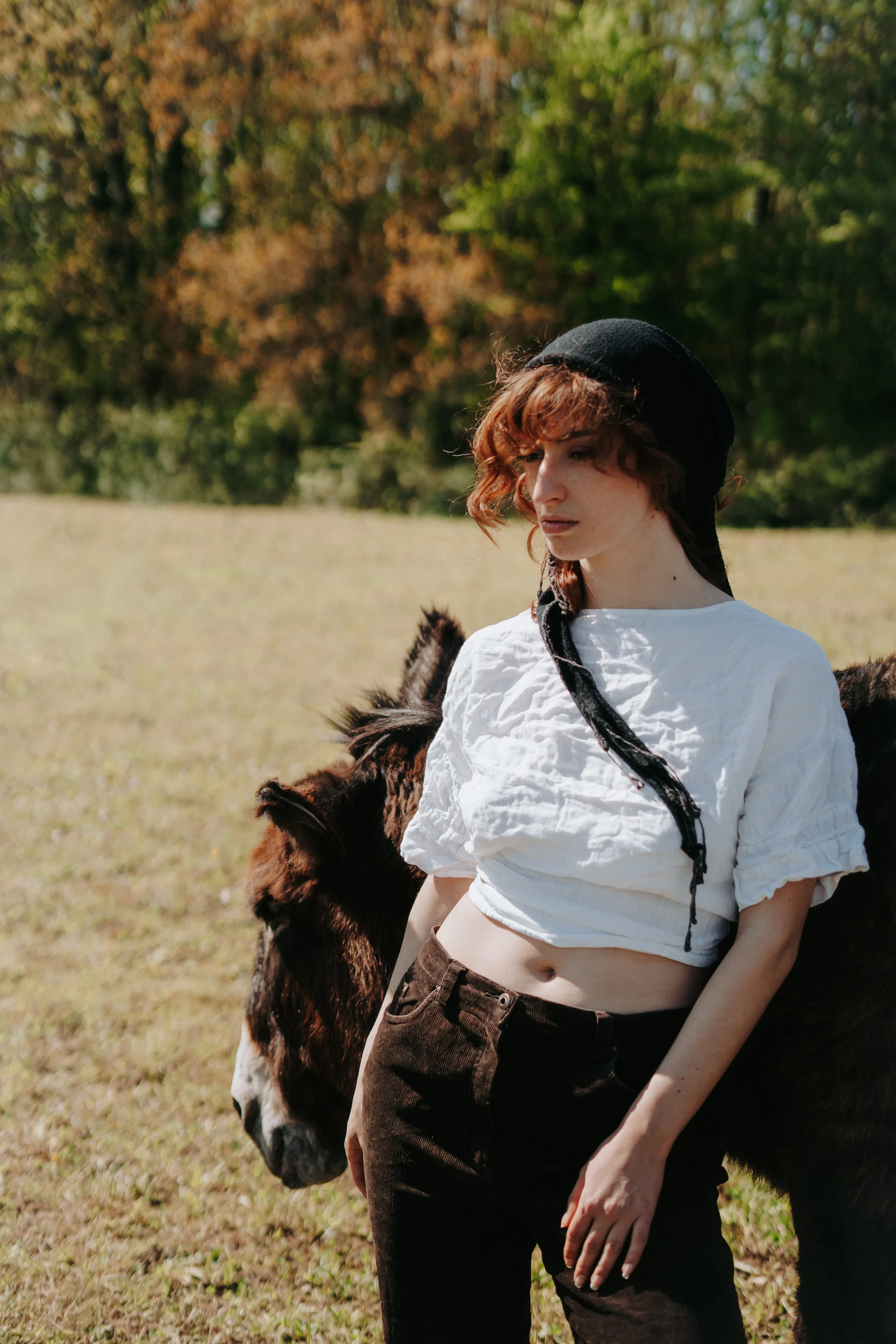 Une jeune femme aux cheveux bouclés roux portant un bonnet noir, une blouse blanche parachutée, et un pantalon marron, debout près d'un cheval dans un champ en plein air avec des arbres en automne en arrière-plan.