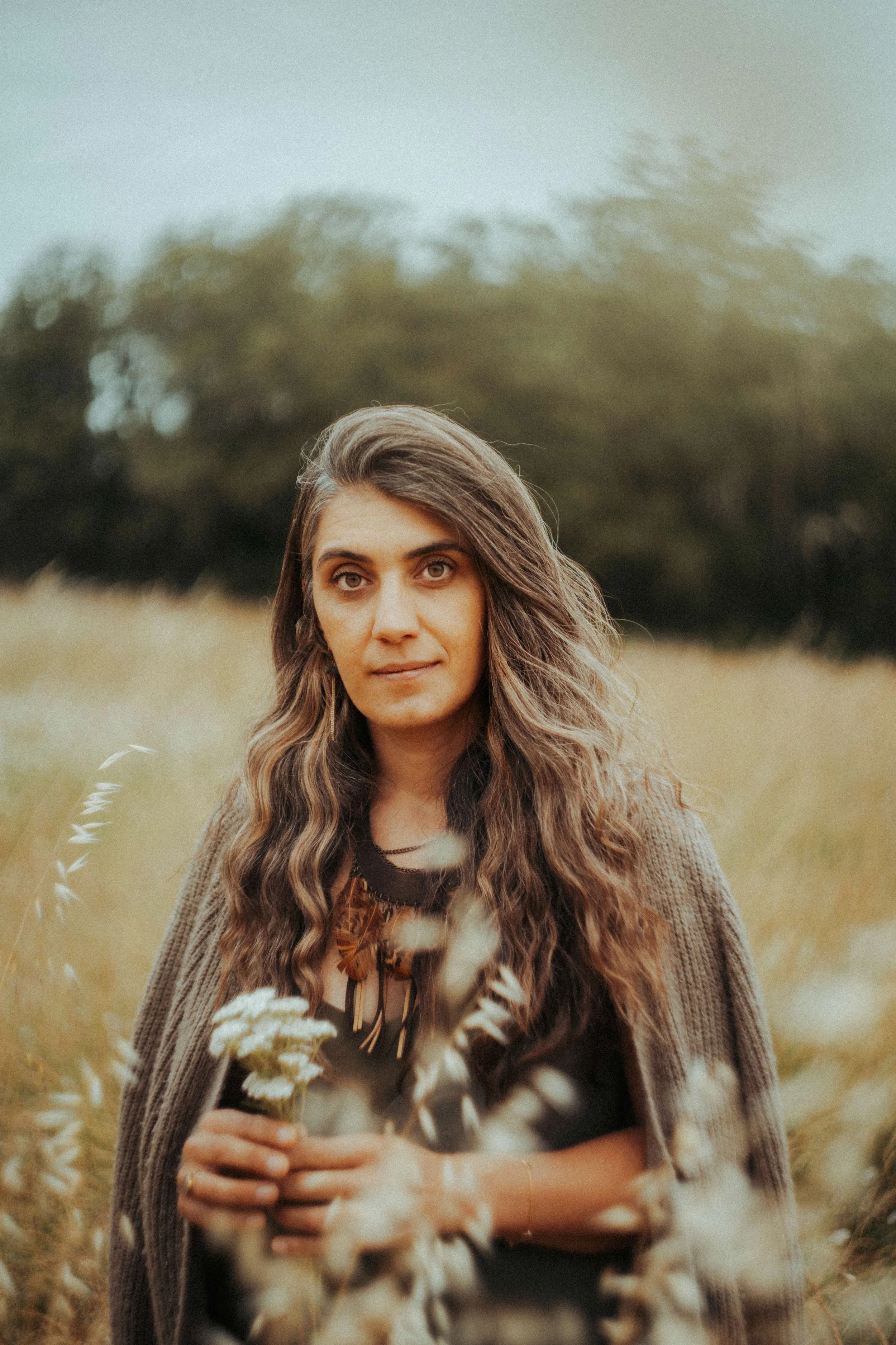 une femme aux longs cheveux ondulés tenant un bouquet de fleurs dans un champ, portant un vêtement d'extérieur marron, avec un arrière-plan de champs et d'arbres, dans une lumière douce d'après-midi.