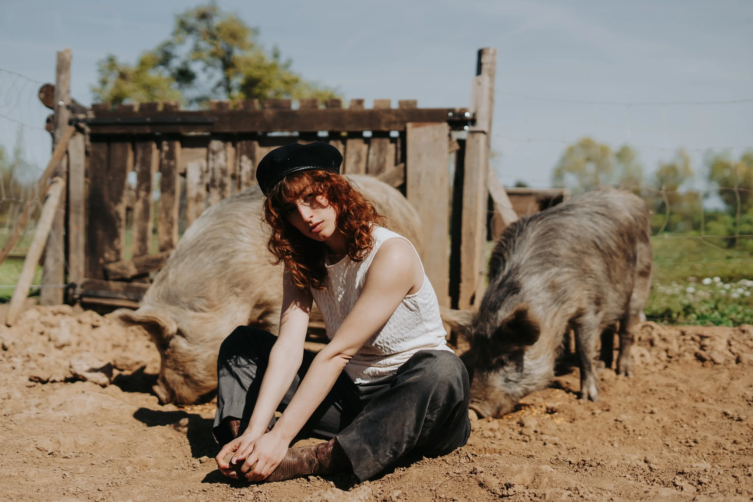 Une femme aux cheveux bouclés rouges, portant un béret noir et une blouse blanche, assise par terre dans une ferme avec des cochons, sous un ciel bleu avec quelques arbres en arrière-plan.
