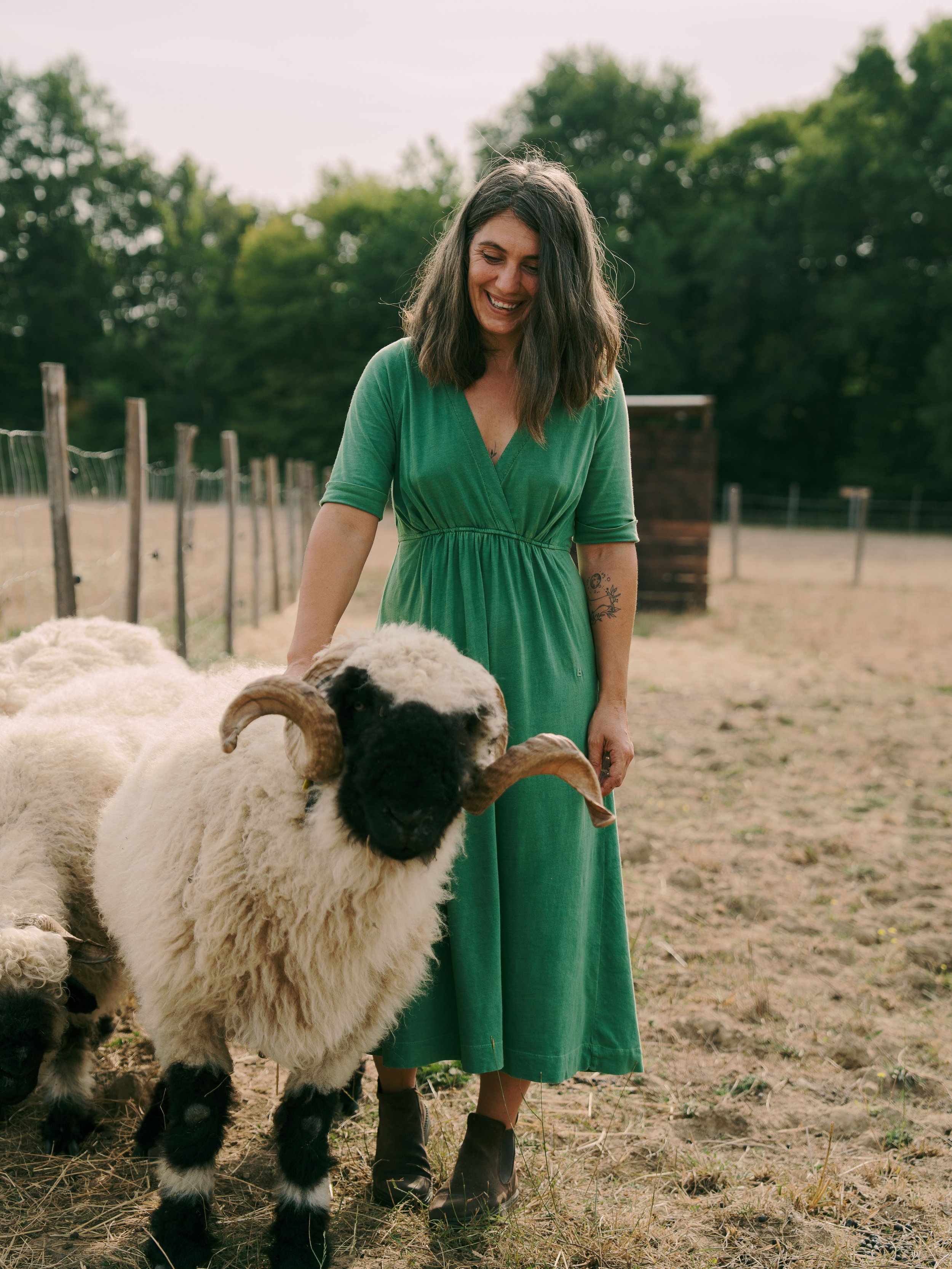 Une femme en robe verte sourit en tenant un mouton avec de grandes cornes dans un pré en extérieur, entourée d'arbres.