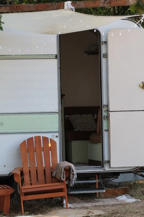 Une caravane avec une porte ouverte, une chaise en bois à l'extérieur, et des guirlandes lumineuses suspendues au-dessus.
