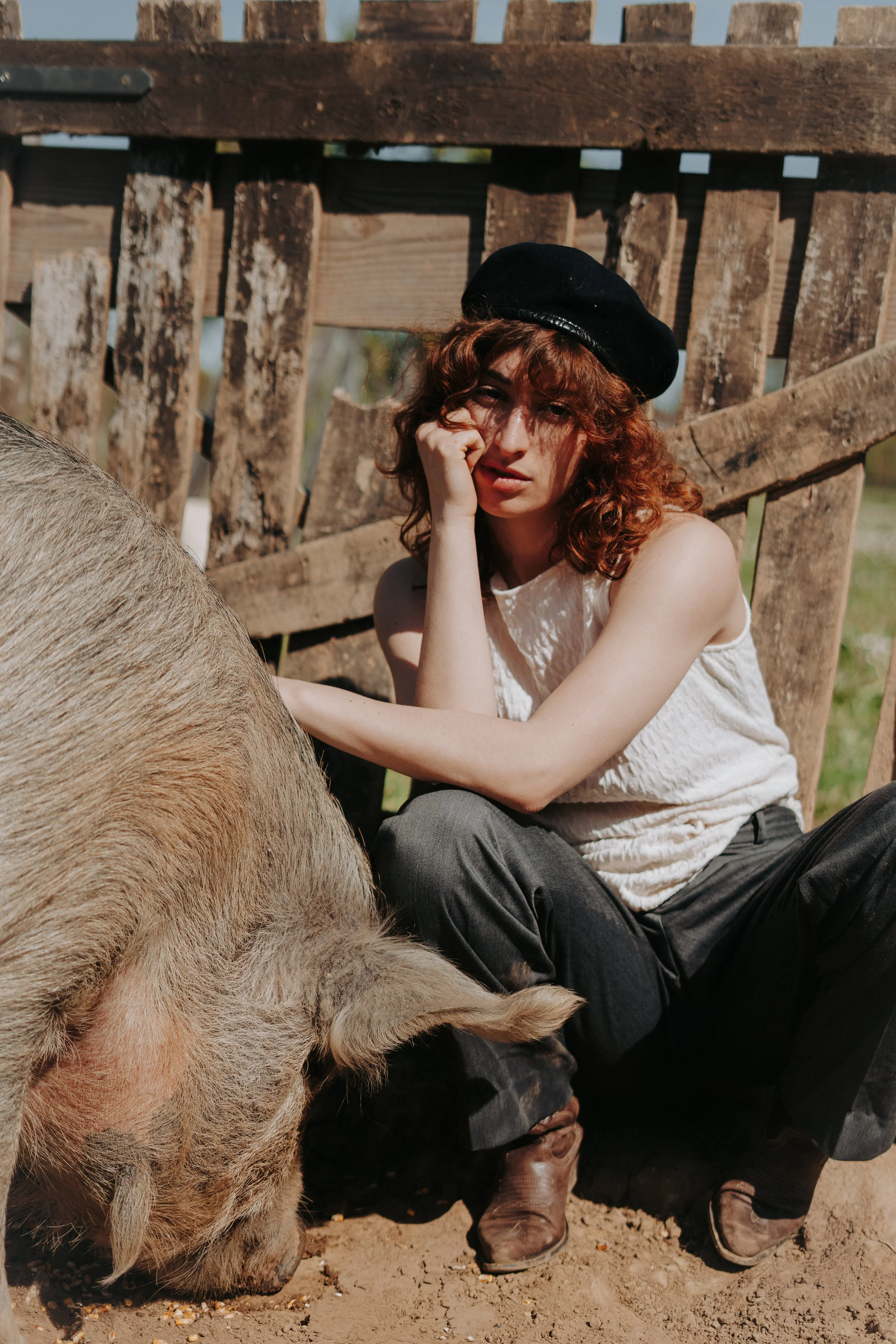 Une jeune femme aux cheveux bouclés rousse portant un béret noir, assise par terre près d'un cochon, dans un environnement extérieur avec une clôture en bois derrière elle.