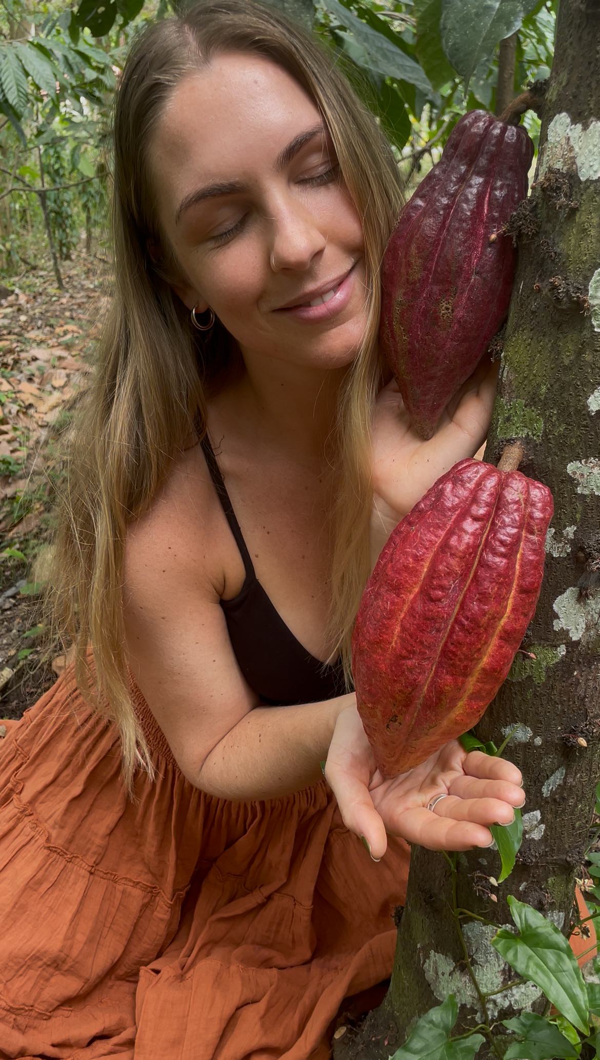 A woman with long hair smiling with her eyes closed, hugging a tree that has large red leaves.