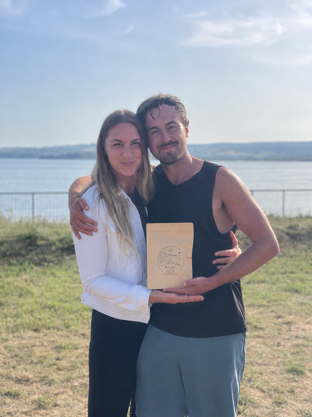 A man and woman stand outdoors near a body of water, hugging and holding a package with a circular logo that reads 'Back Self,' both smiling at the camera.