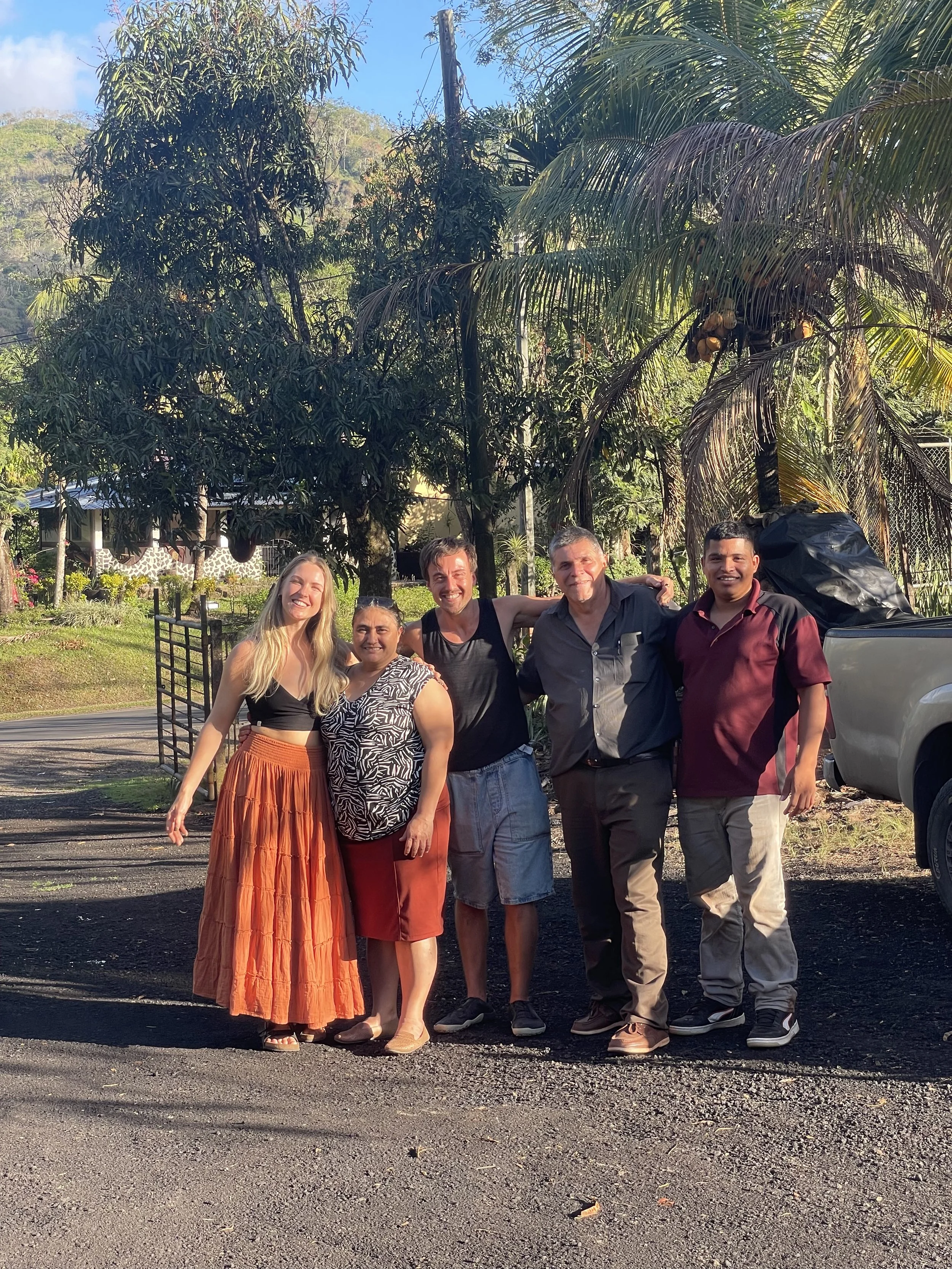 A group of six people, four men and two women, standing outdoors with trees and a house in the background, smiling for the camera.