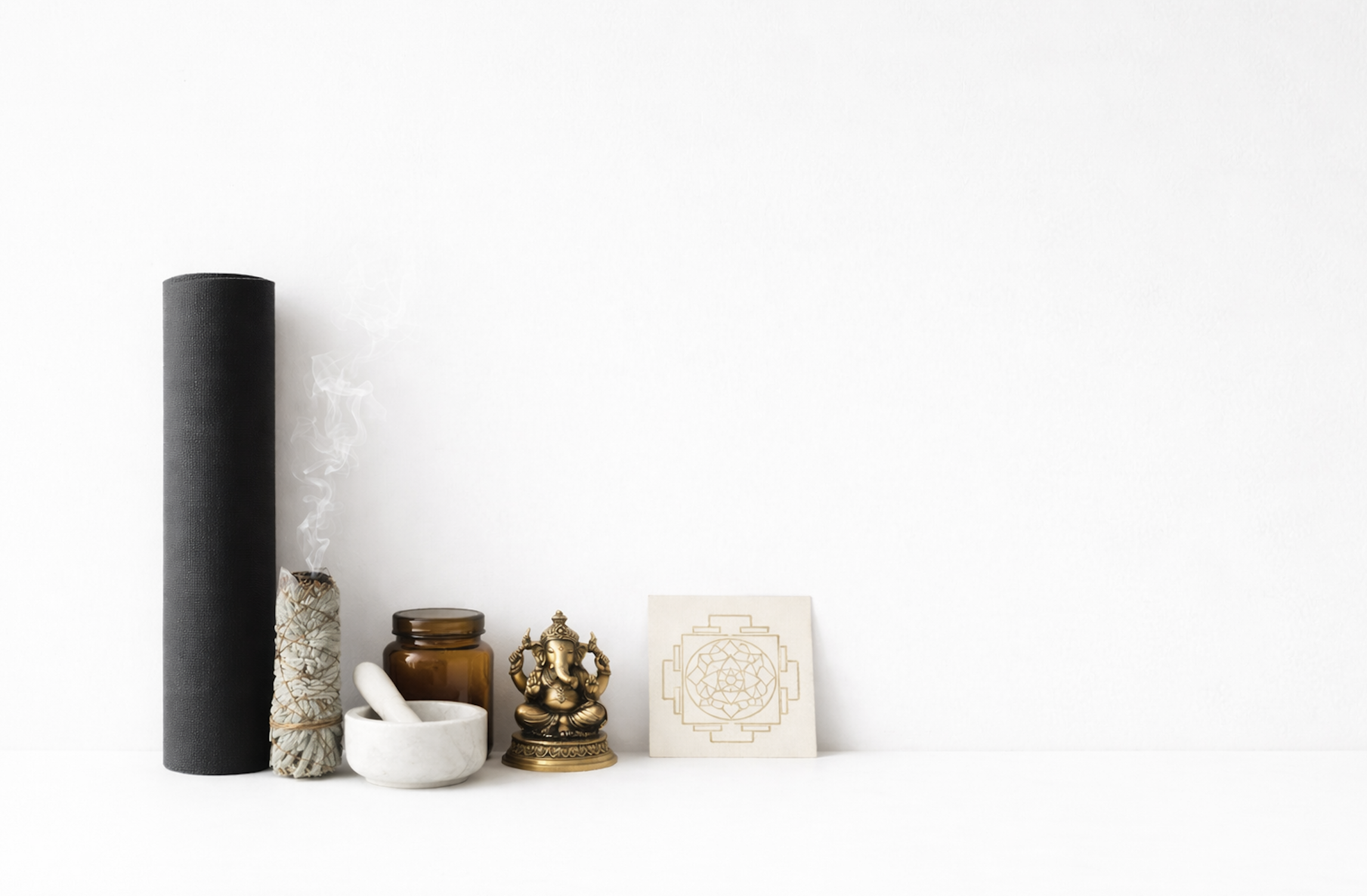 A collection of spiritual and wellness objects arranged on a white surface against a white wall, including a black candle, a depiction of white sage, a small mortar and pestle, a brown glass jar, a brass Ganesha statue, and spiritual artwork.