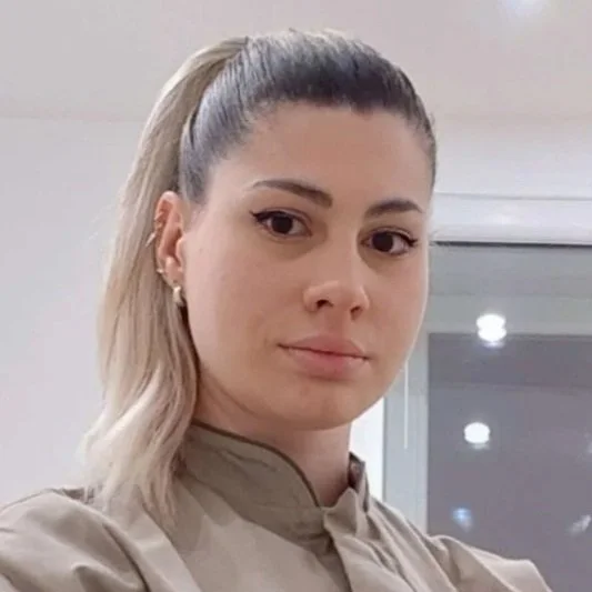 Jeune femme avec cheveux blonds attachés en queue de cheval, portant une chemise beige, regardant l'appareil photo.