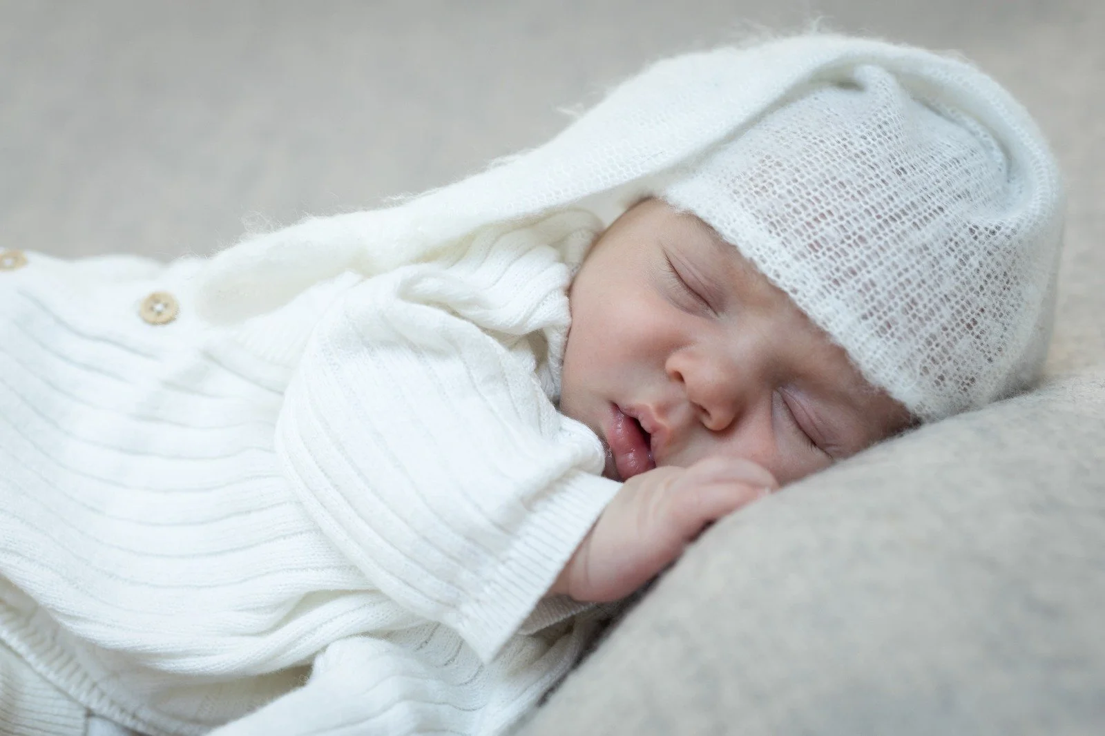 Un bébé dormant paisiblement, portant un bonnet blanc en tricot et habillé d'un vêtement blanc avec boutons, allongé sur une surface douce.