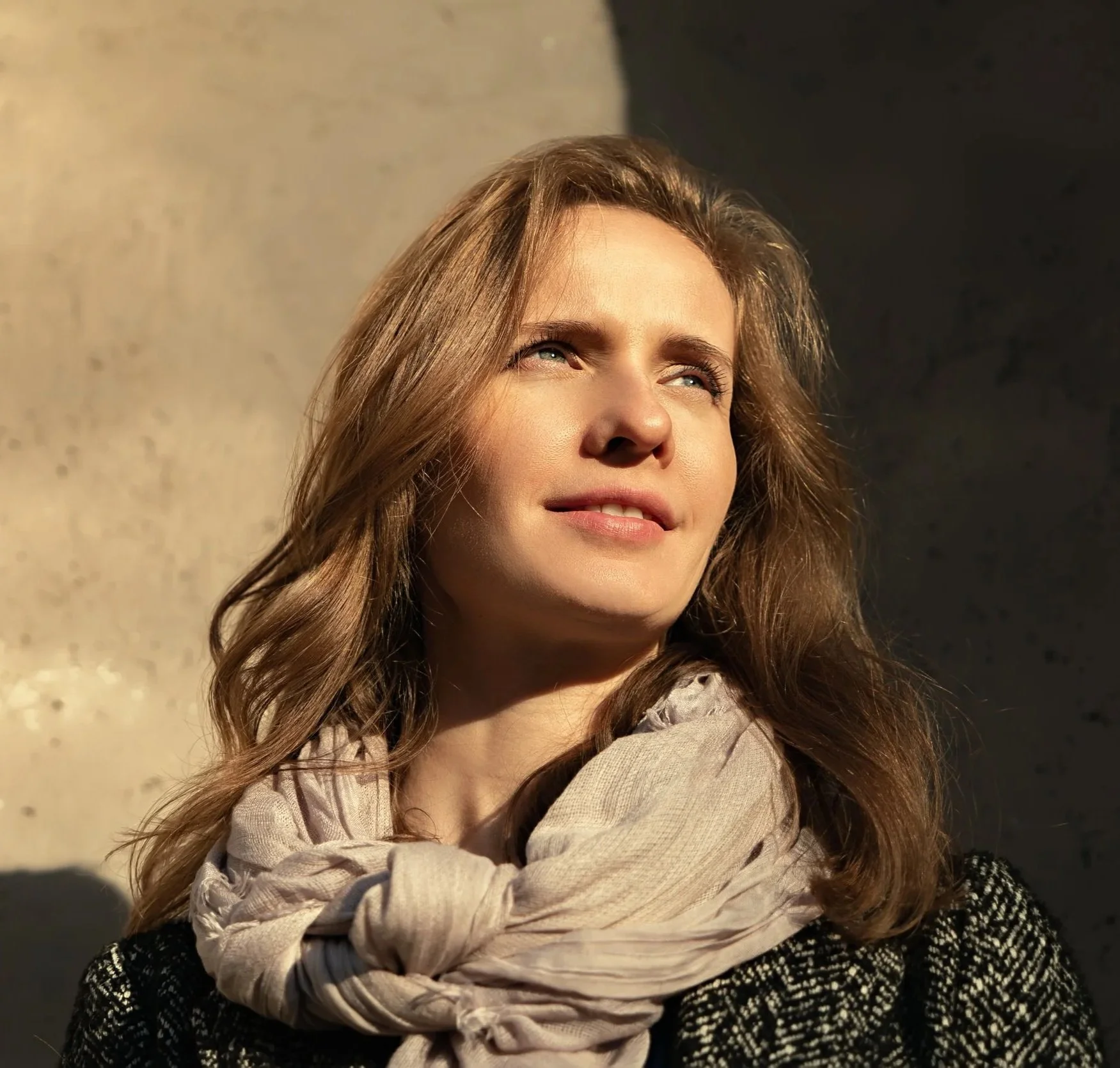 Une femme aux cheveux longs et bouclés, portant une écharpe beige et un pull noir à motif blanc, regarde vers le haut sous un ciel ensoleillé.